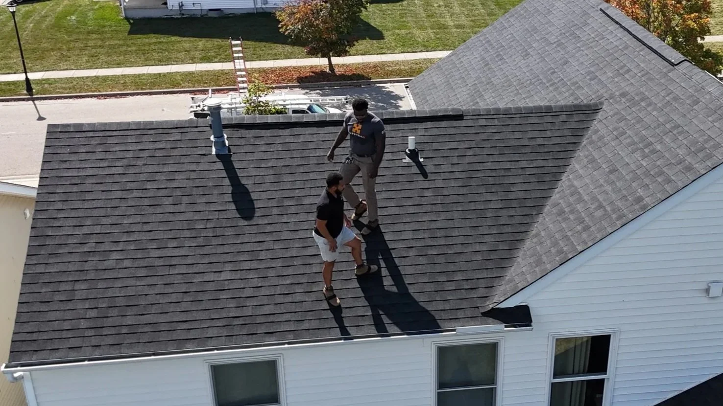 Two men working on a house roof during daytime; one standing and one kneeling.