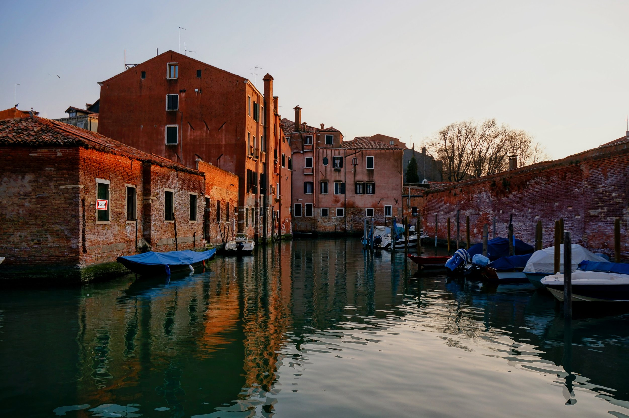 Photo taken by Christina Sarelakos of Venetian canal with boats, brick buildings, and a warm sunset glow.