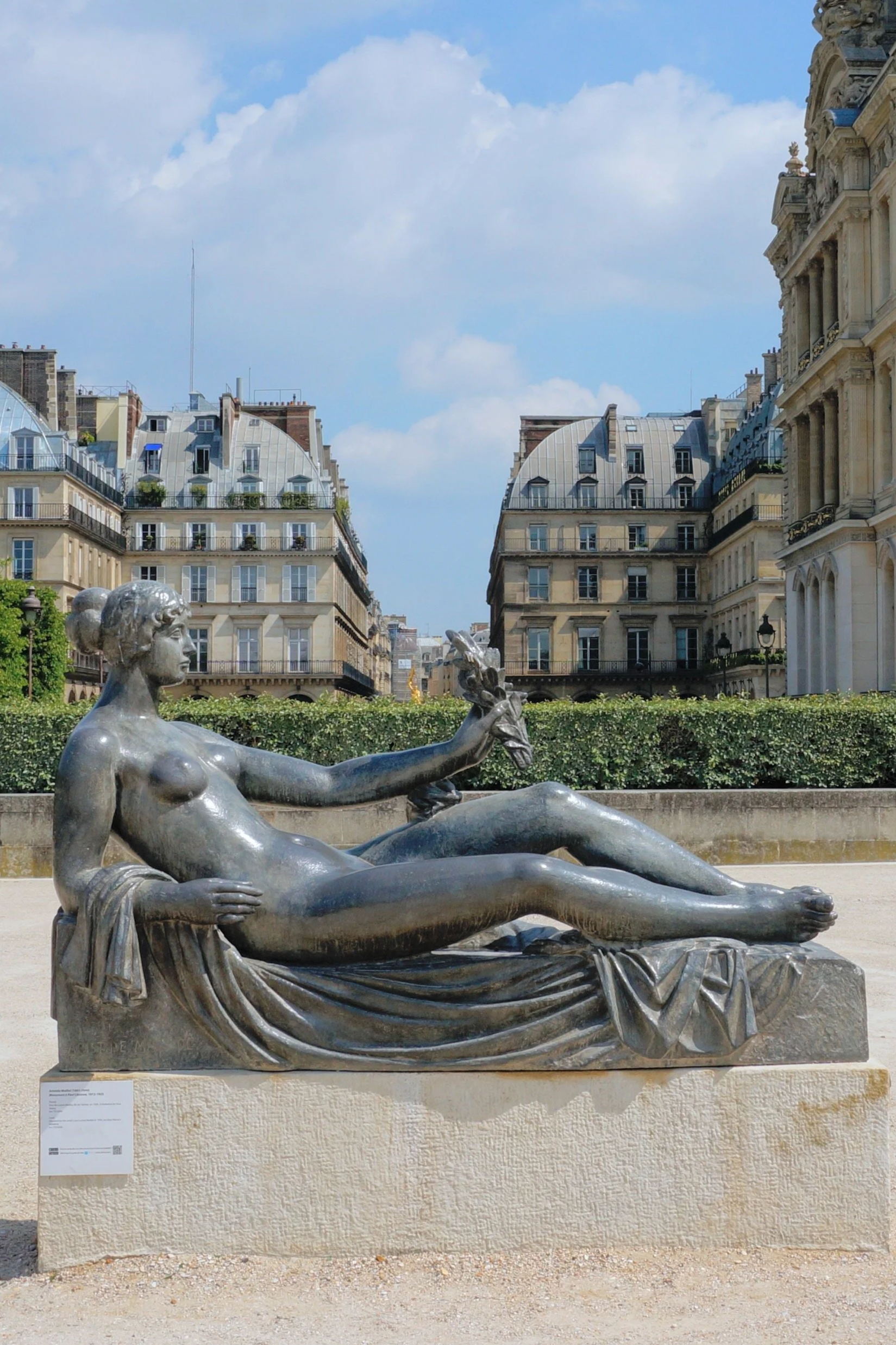 Reclining bronze statue of a nude figure holding a small object, set against historic buildings and blue sky.