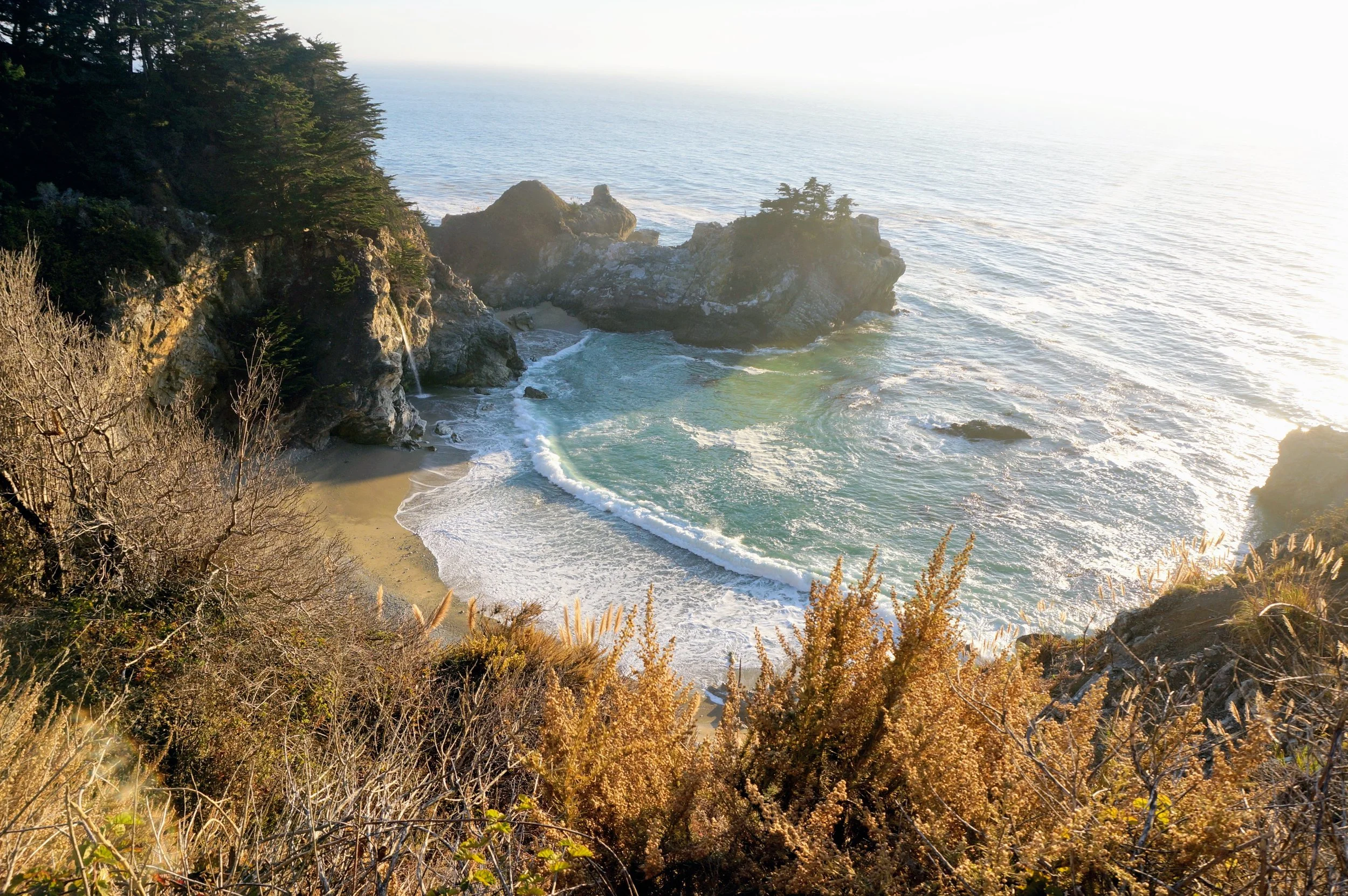 Coastal view with cliffs, ocean waves, and sandy beach at McWay Falls, Big Sur, California.