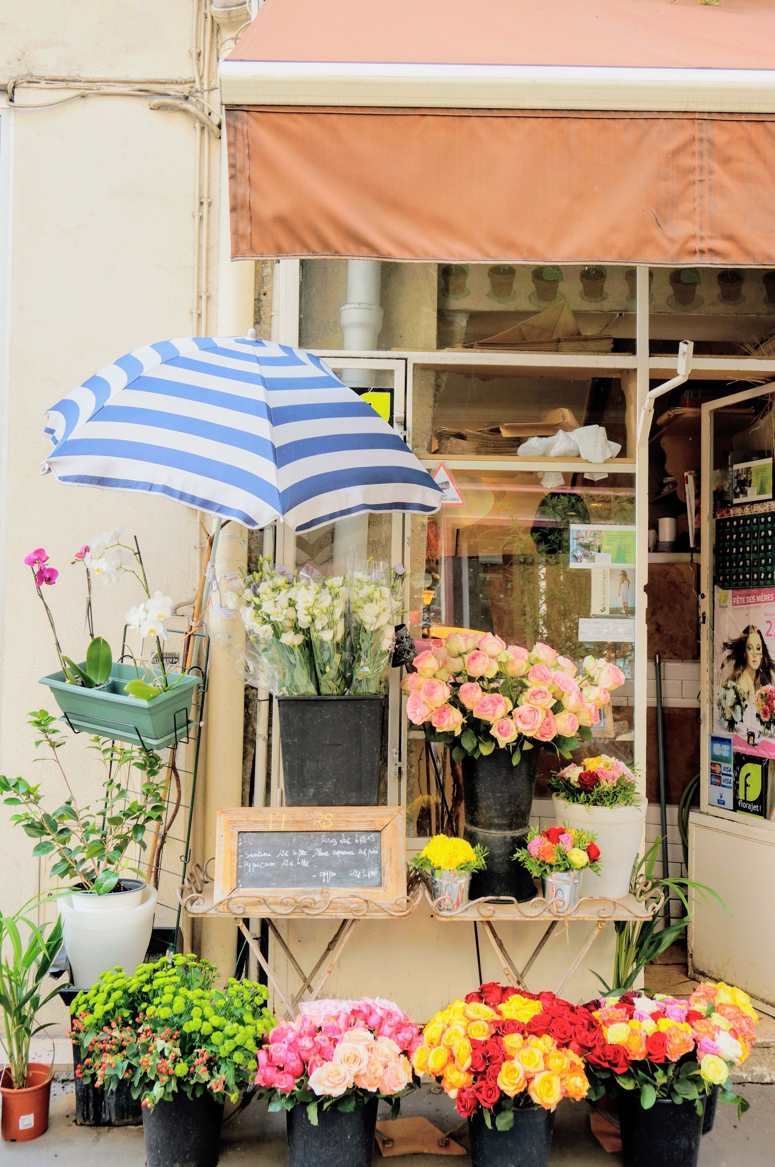Flower shop exterior with colorful flowers under a striped umbrella and brown awning.