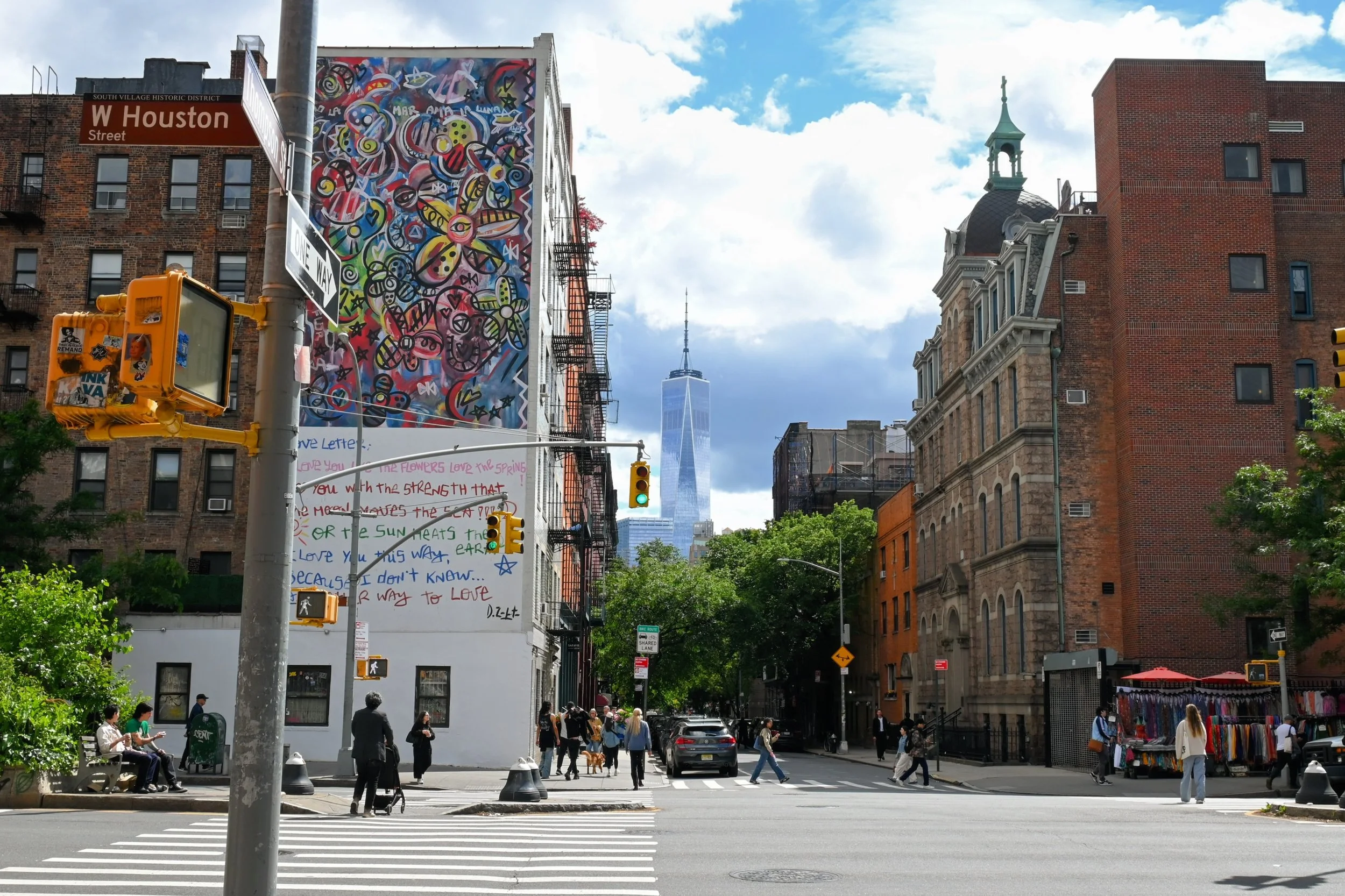 City street scene in New York City with pedestrians crossing at a crosswalk; One World Trade Center in the background; colorful murals on buildings; street signs and traffic lights; trees lining the street; vendor stalls with clothing.
