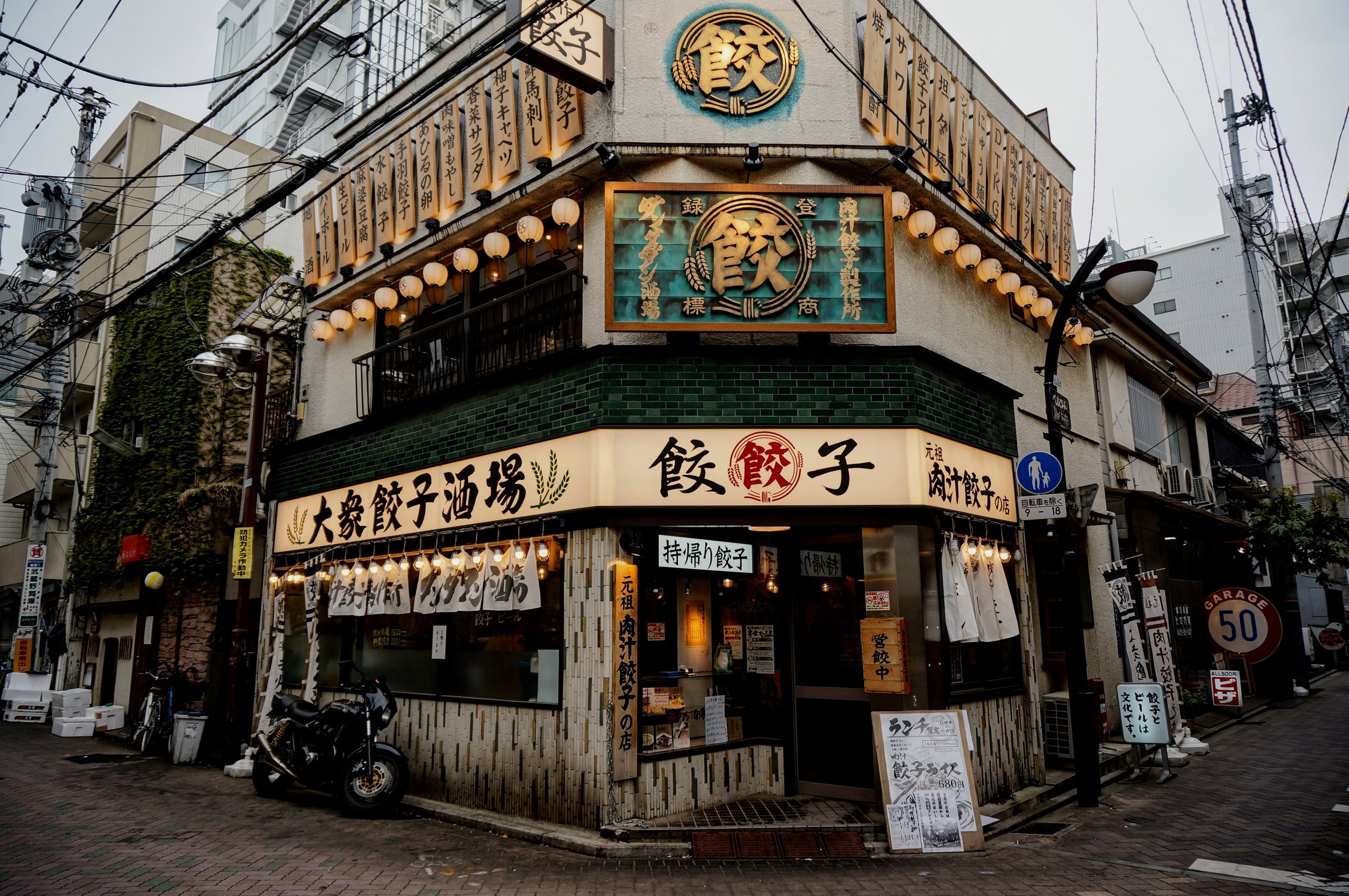 Japanese gyoza restaurant with neon signs and outdoor lights on a city street corner, featuring a motorcycle parked outside and urban buildings in the background.
