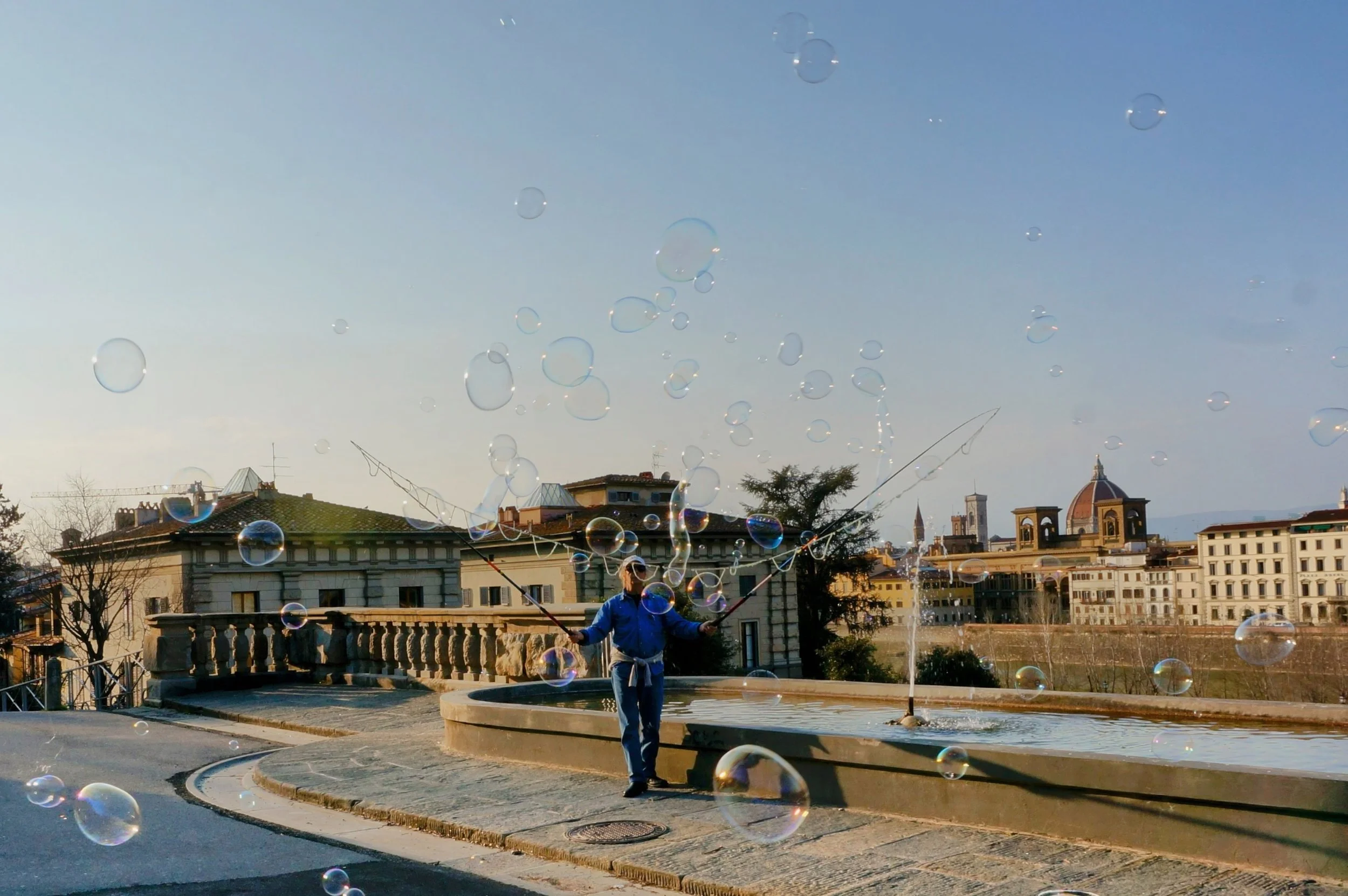 Person making bubbles outdoors near a fountain, with an urban skyline in the background under a clear sky.