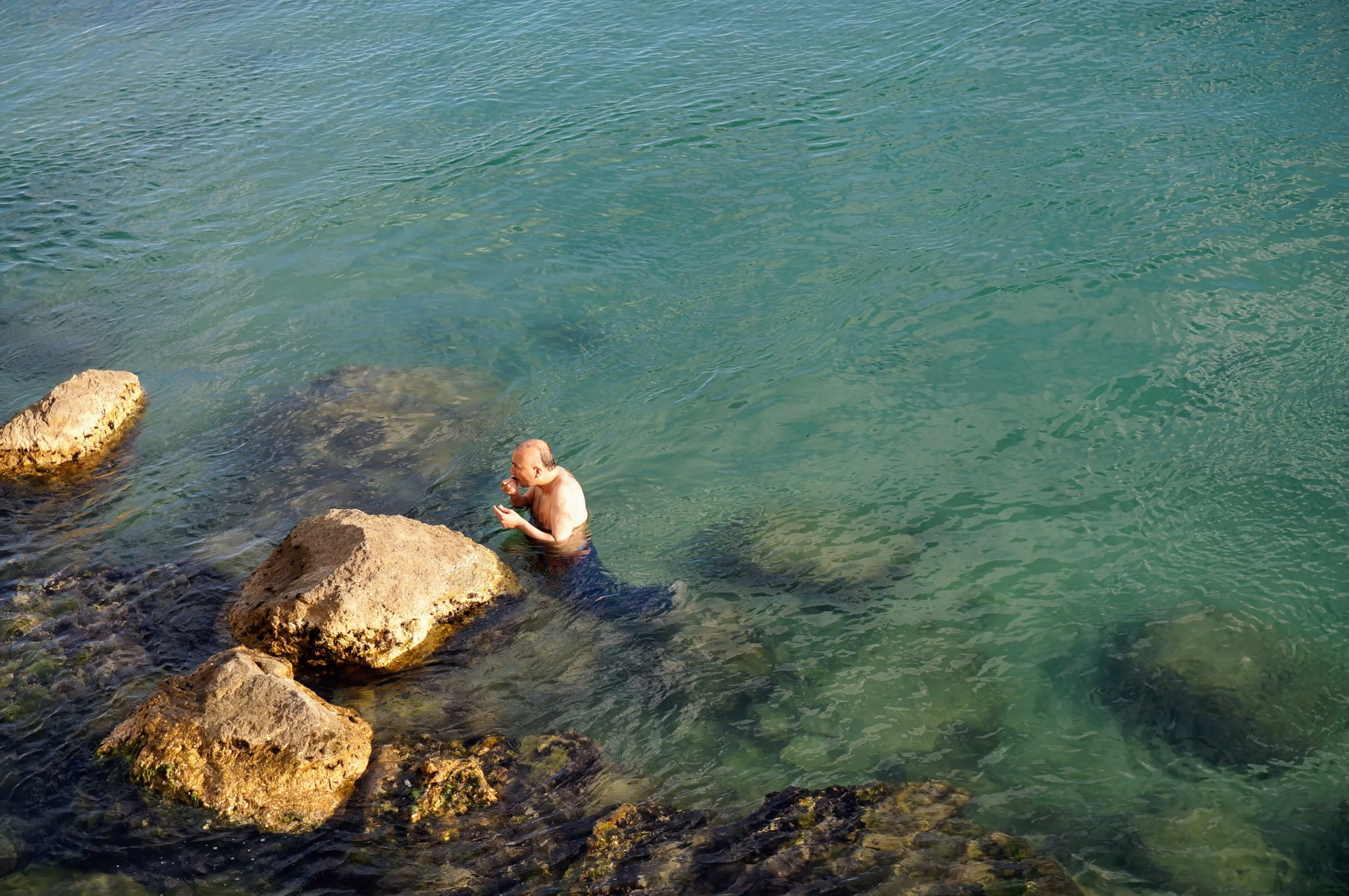 Man standing in shallow, clear water near rocks, washing his face.
