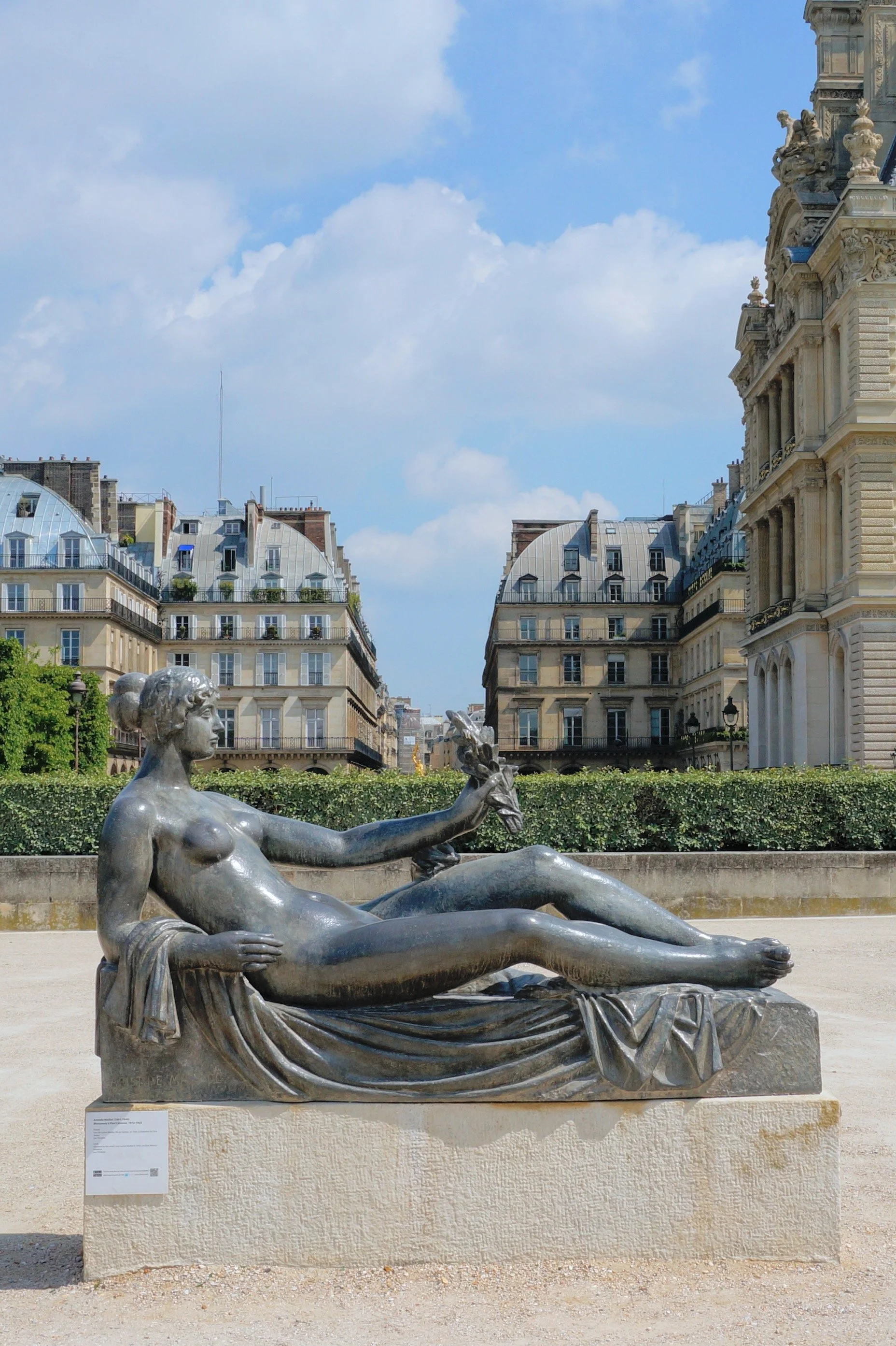 Reclining bronze statue of a nude figure holding a small object, set against historic buildings and blue sky.