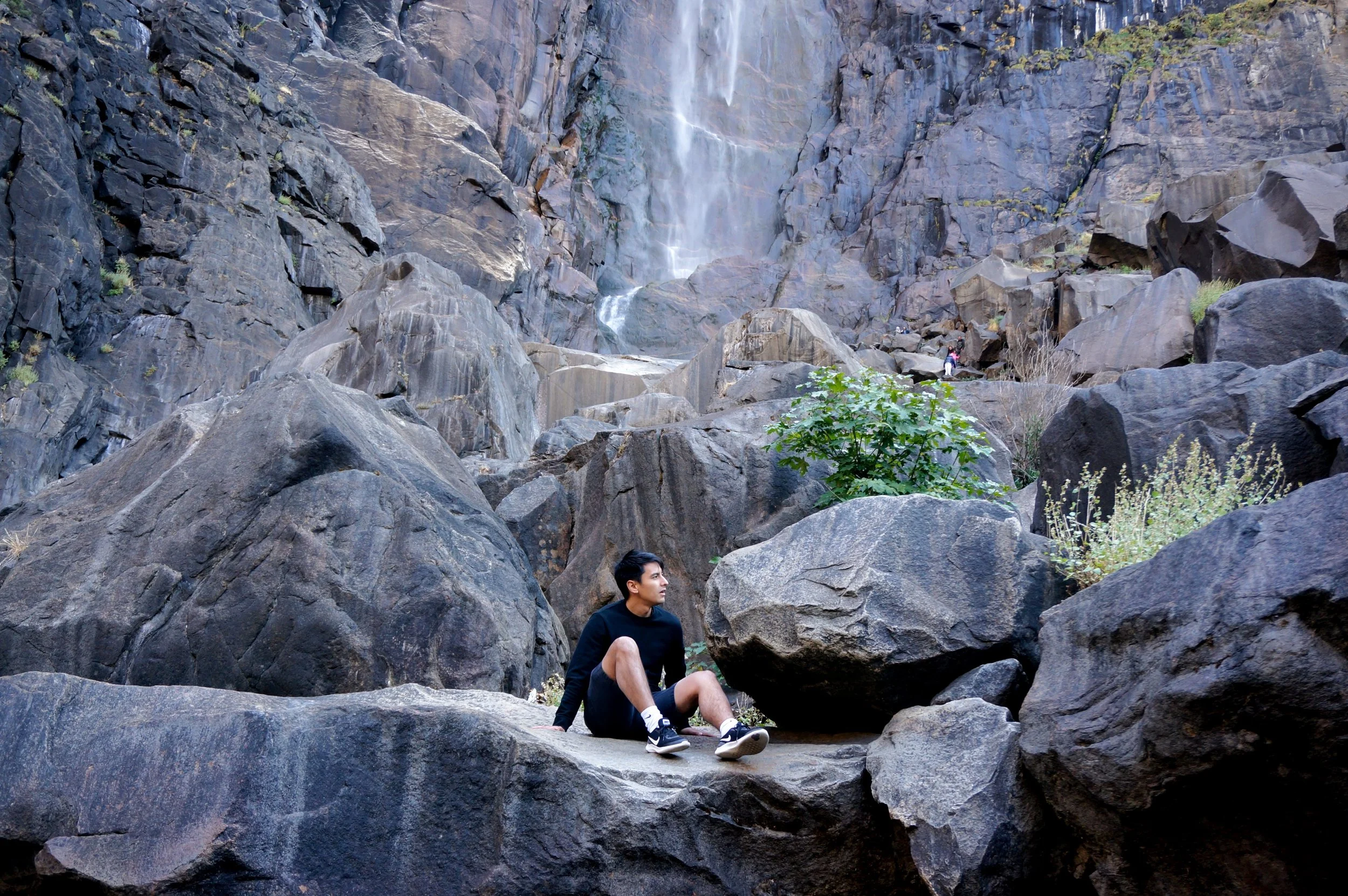 Person sitting on rocks near a waterfall in a rocky canyon.