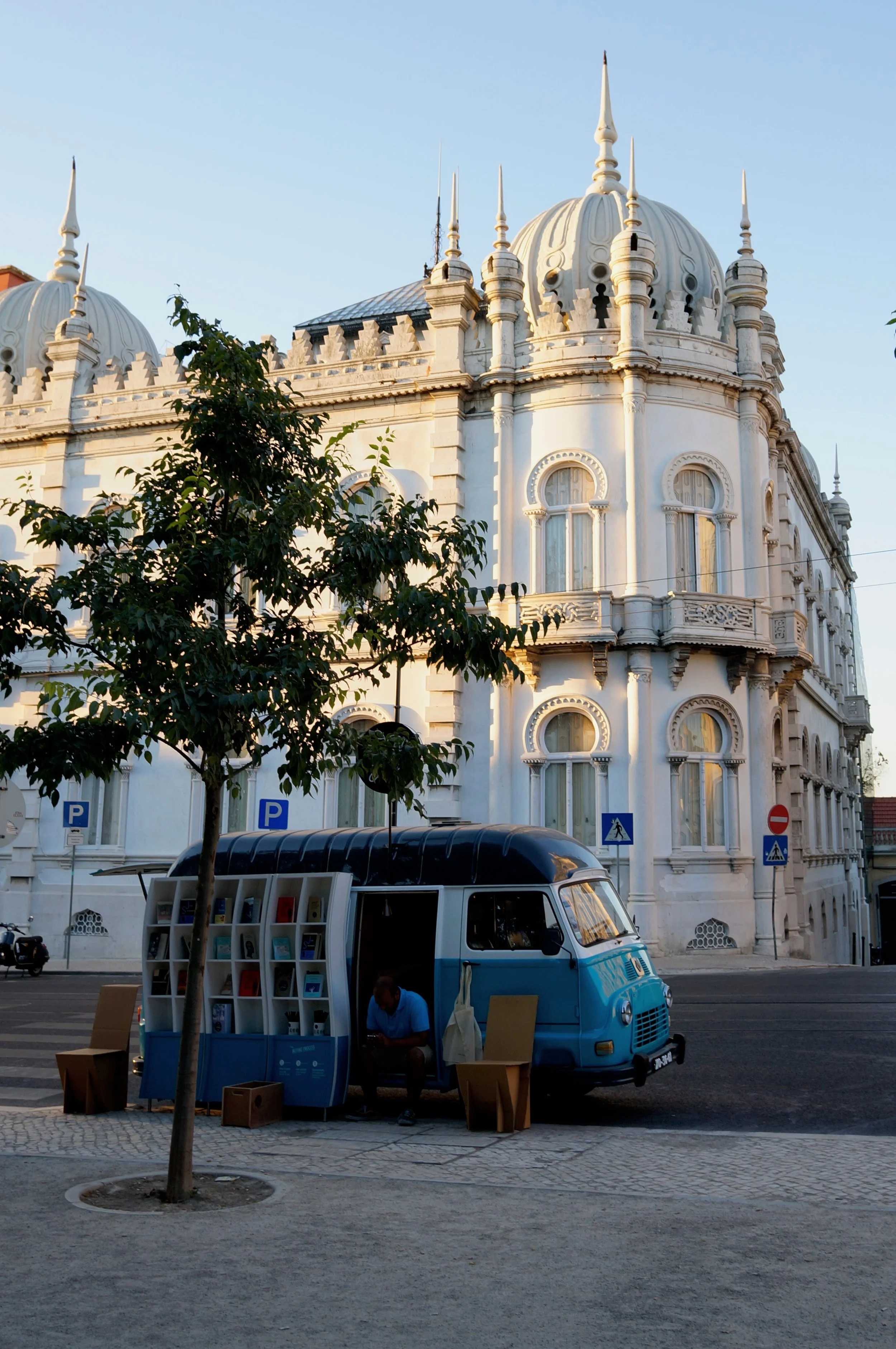 Photo taken by Christina Sarelakos of blue vintage van converted into a bookshop, parked on a street next to a historic white building with ornate architecture in Portugal