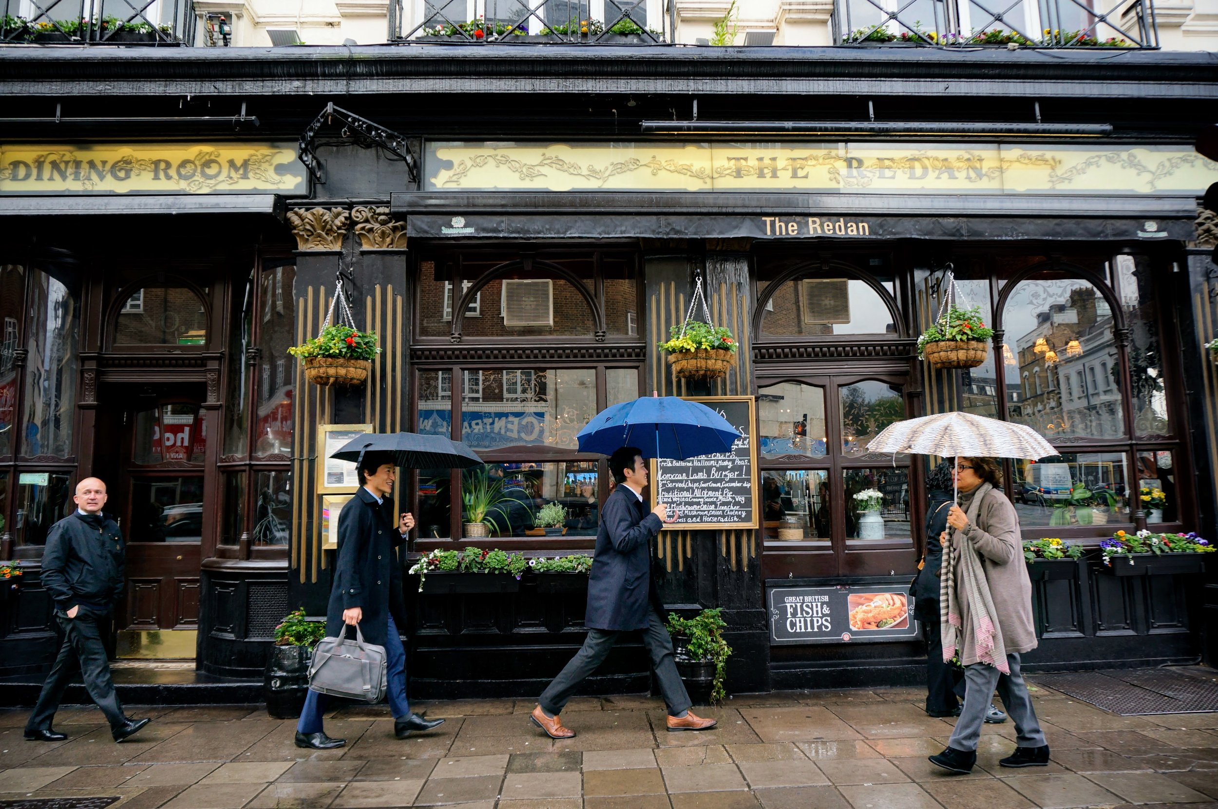 Photo taken by Christina Sarelakos of people walking with umbrellas outside The Redan pub on a rainy street in London