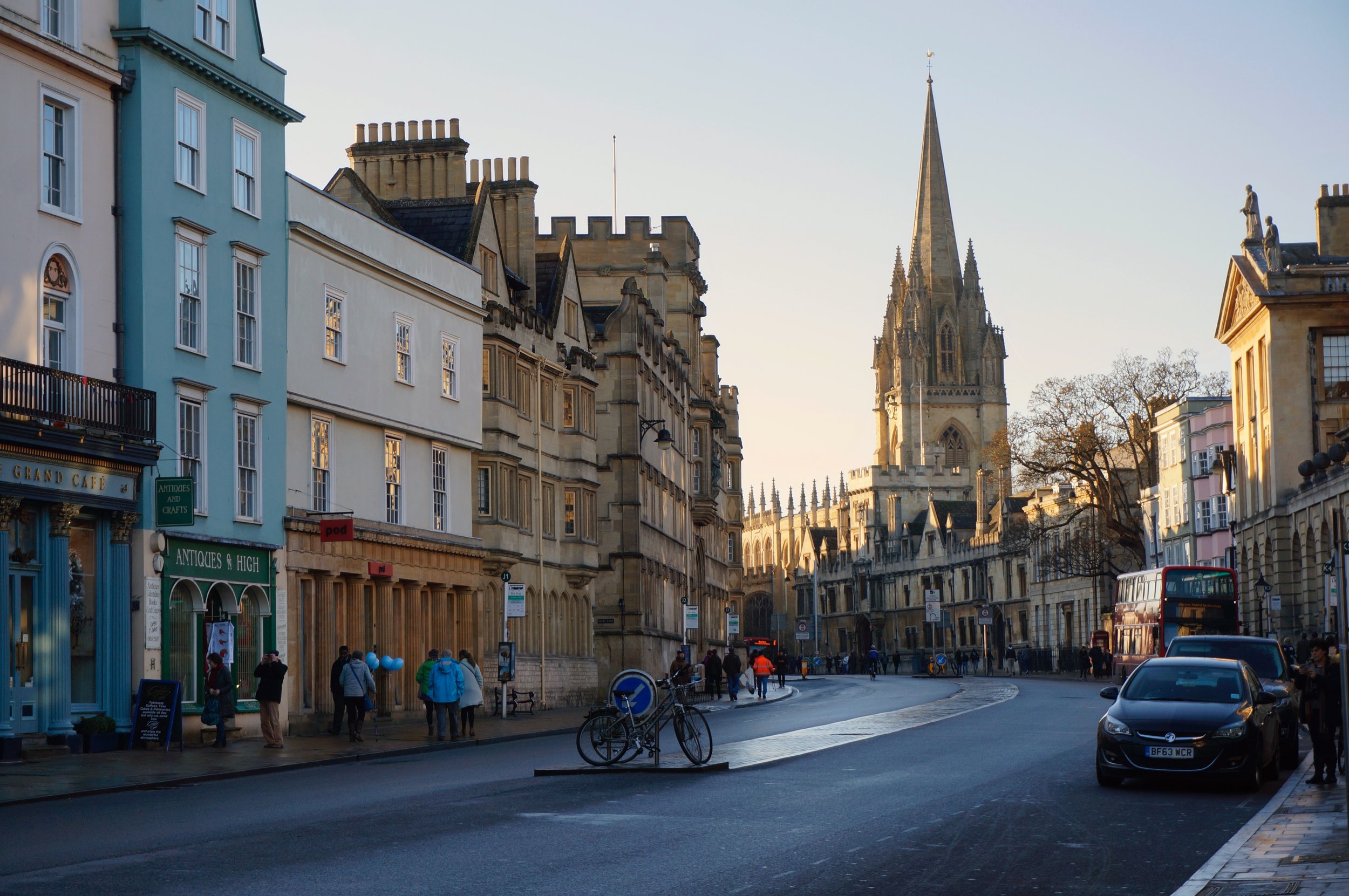 Quaint street scene in Oxford, England with historic buildings, a church spire, and pedestrians on a clear day.