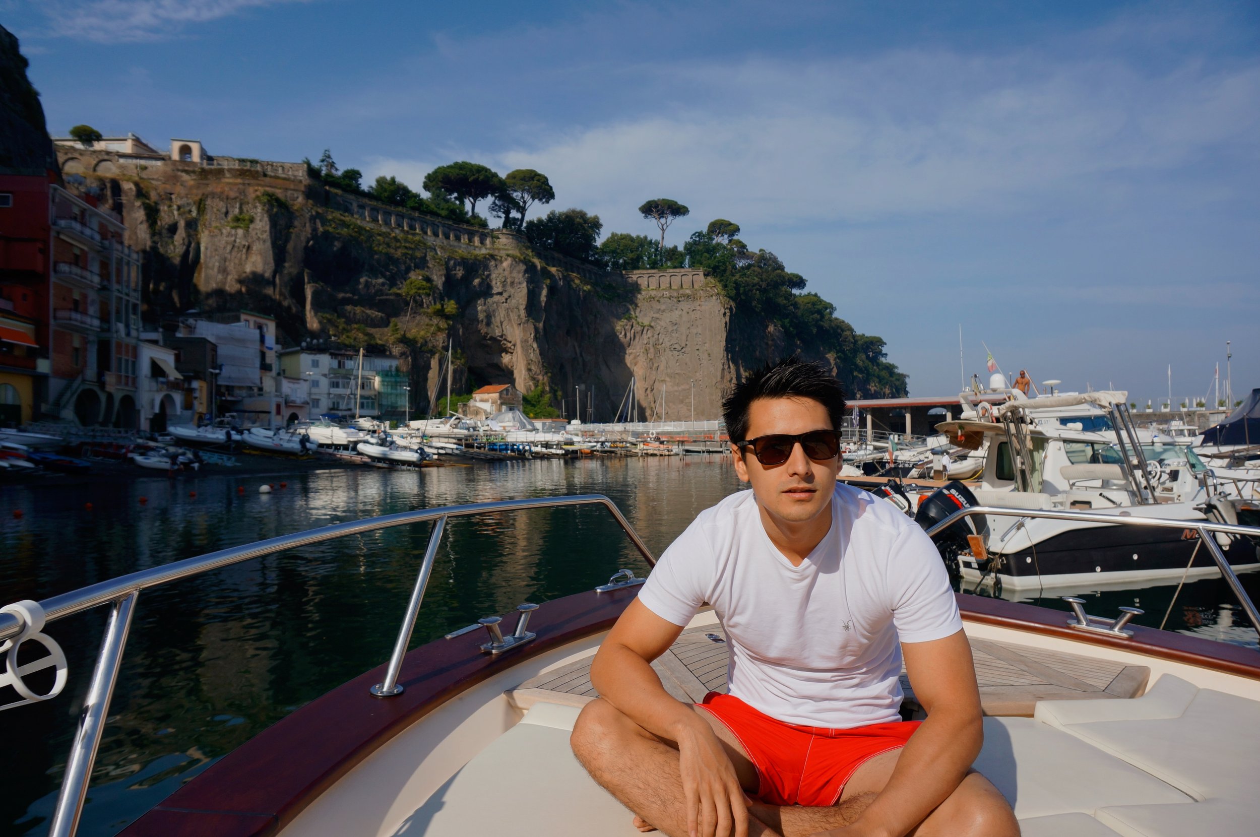 Man wearing sunglasses and red shorts sitting on a boat; harbor with boats and cliffs in the background.