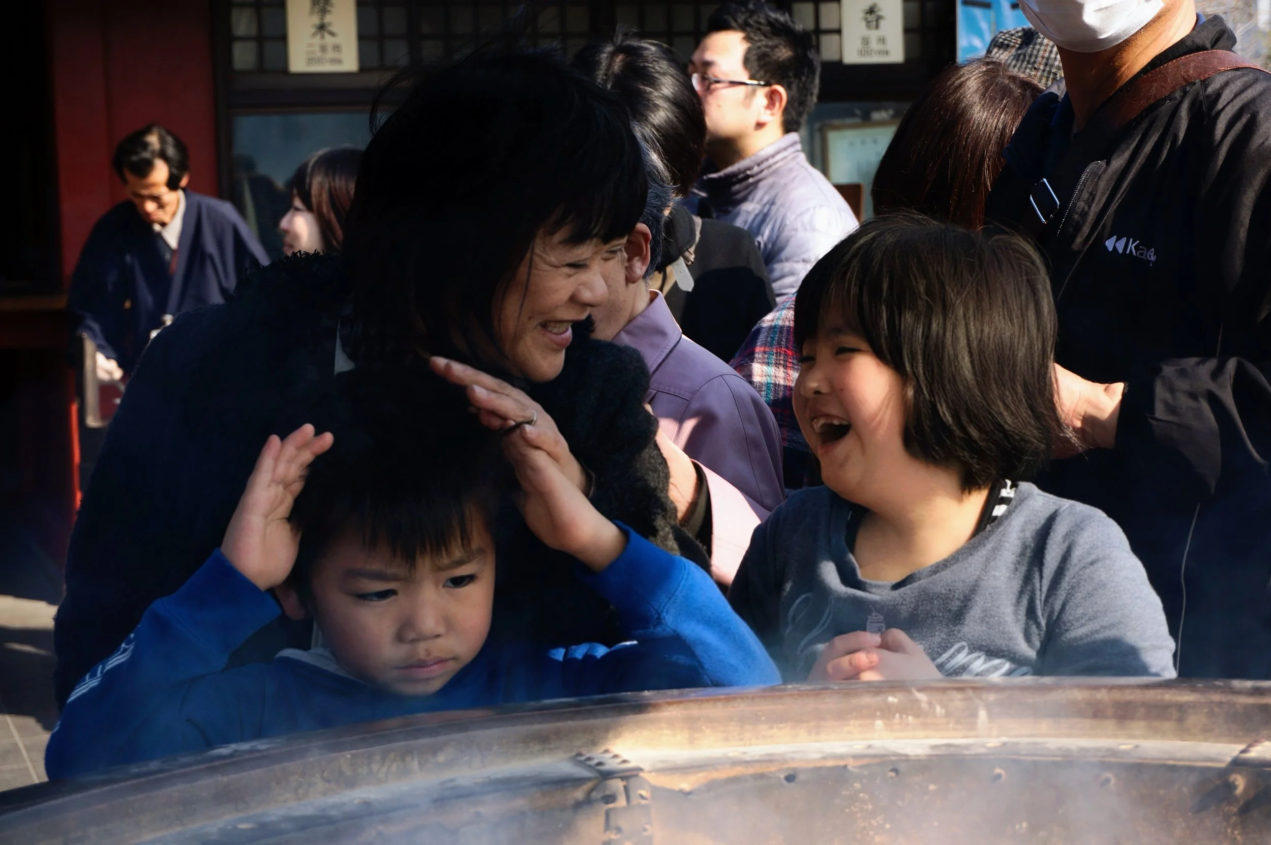 A woman with two children smiling and interacting at a public place, surrounded by a group of people. One child looks serious while the other is laughing. There is a large round object in the foreground with steam or smoke rising from it.