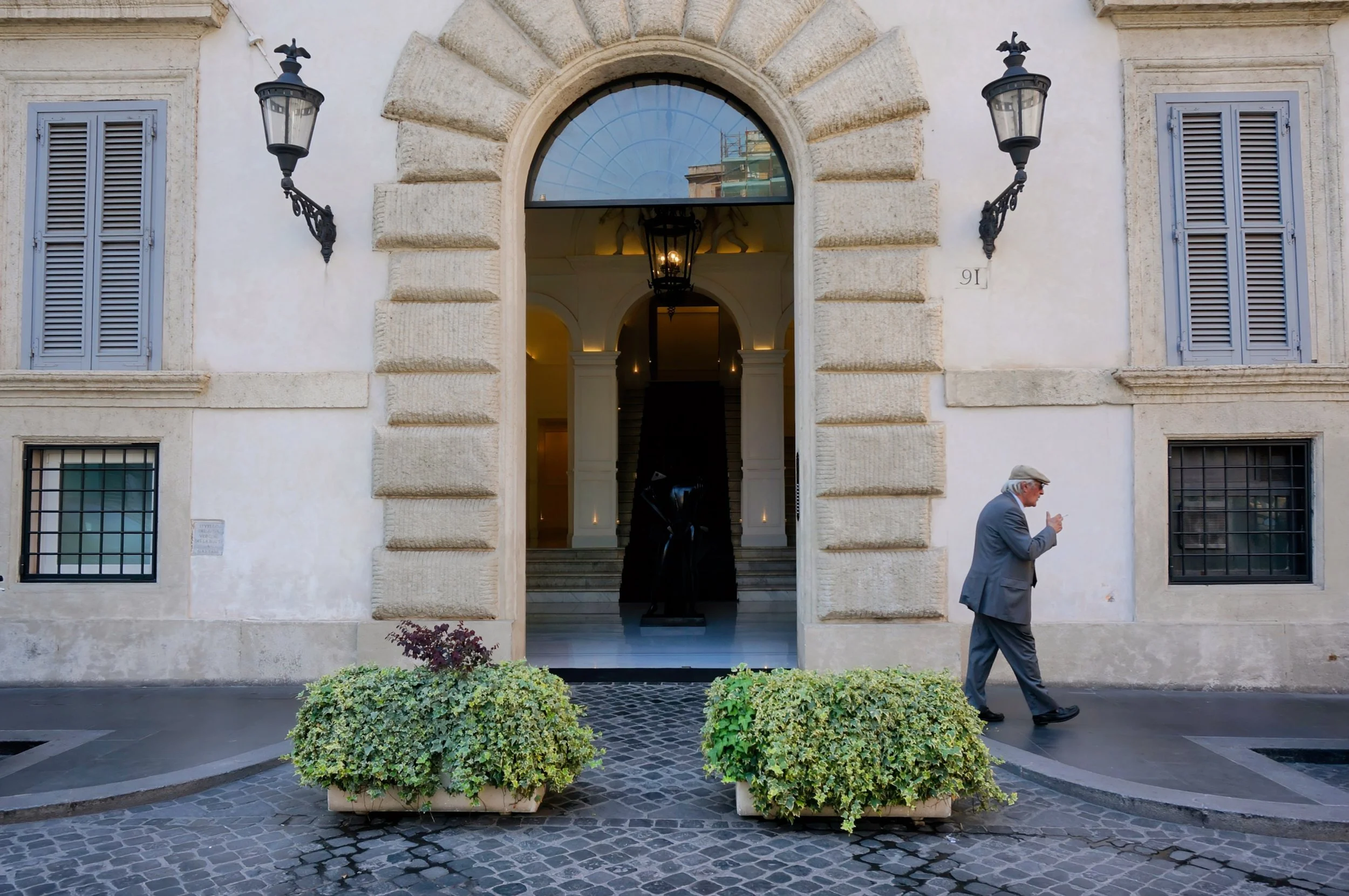 Photo by Christina Sarelakos of elegant arched doorway of a historic building with a man walking by, exterior walls feature decorative stonework and vintage lamps, planters with greenery in front.