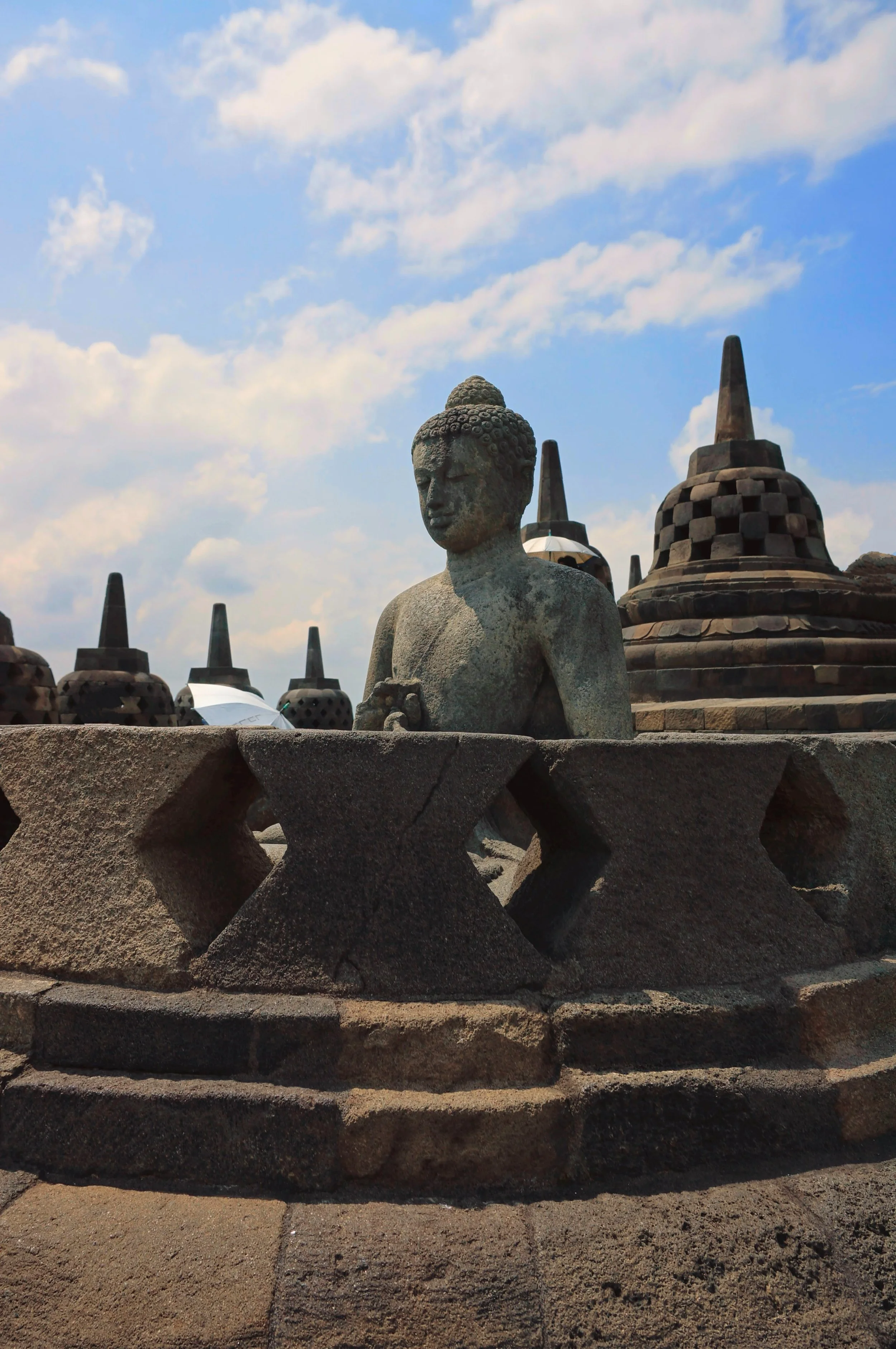 Buddha statue at Borobudur Temple with stupas in the background under a partly cloudy sky.