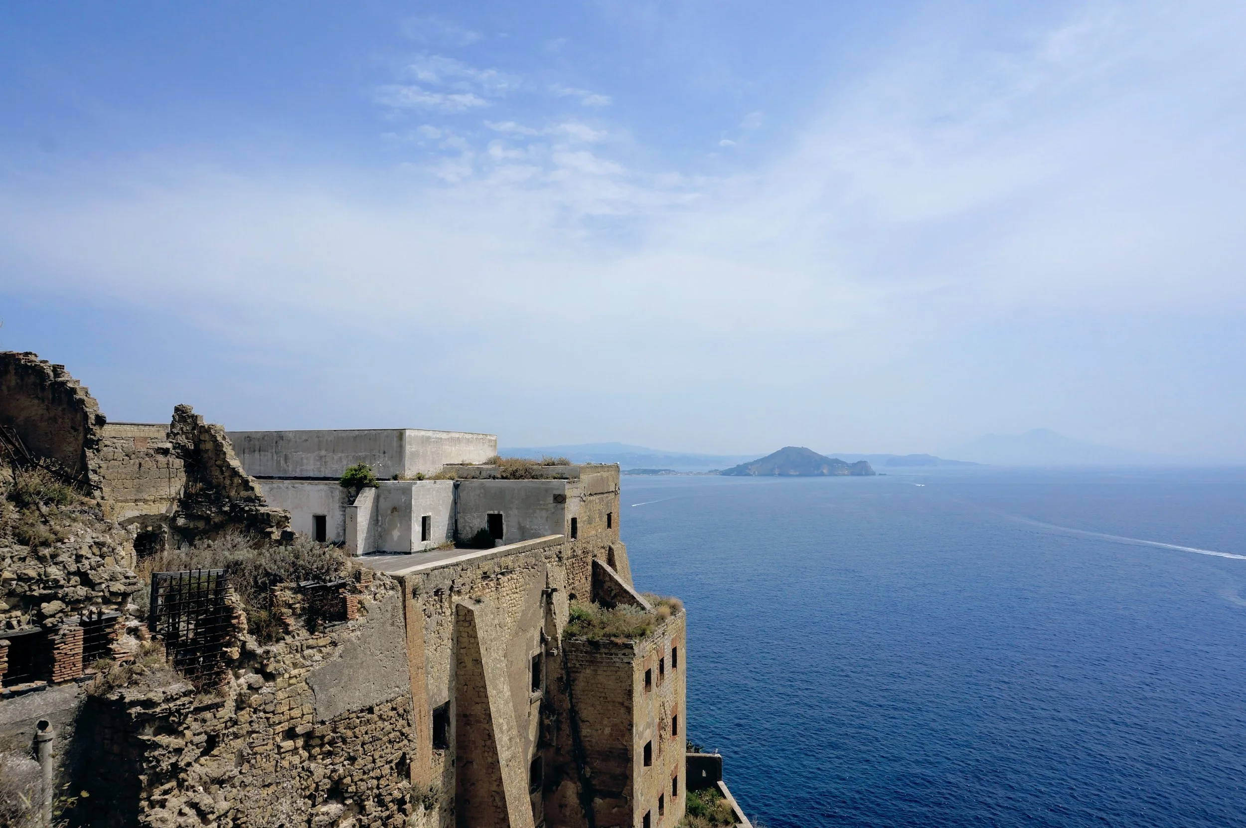 Coastal view with ancient stone fortress ruins and distant islands across a calm sea under a blue sky.