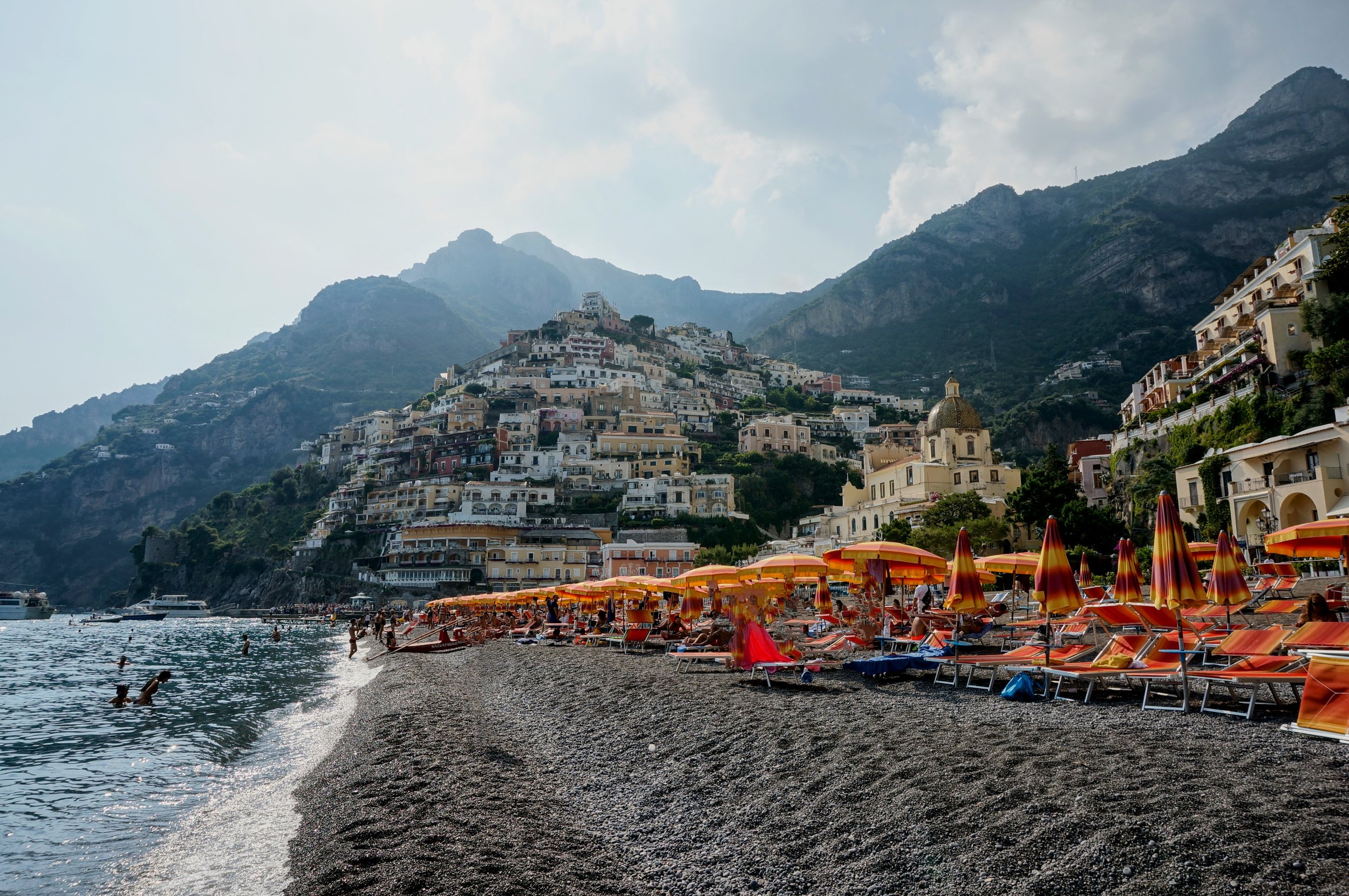 Scenic view of a coastal town with colorful umbrella beach, hillside buildings, and mountains in the background.