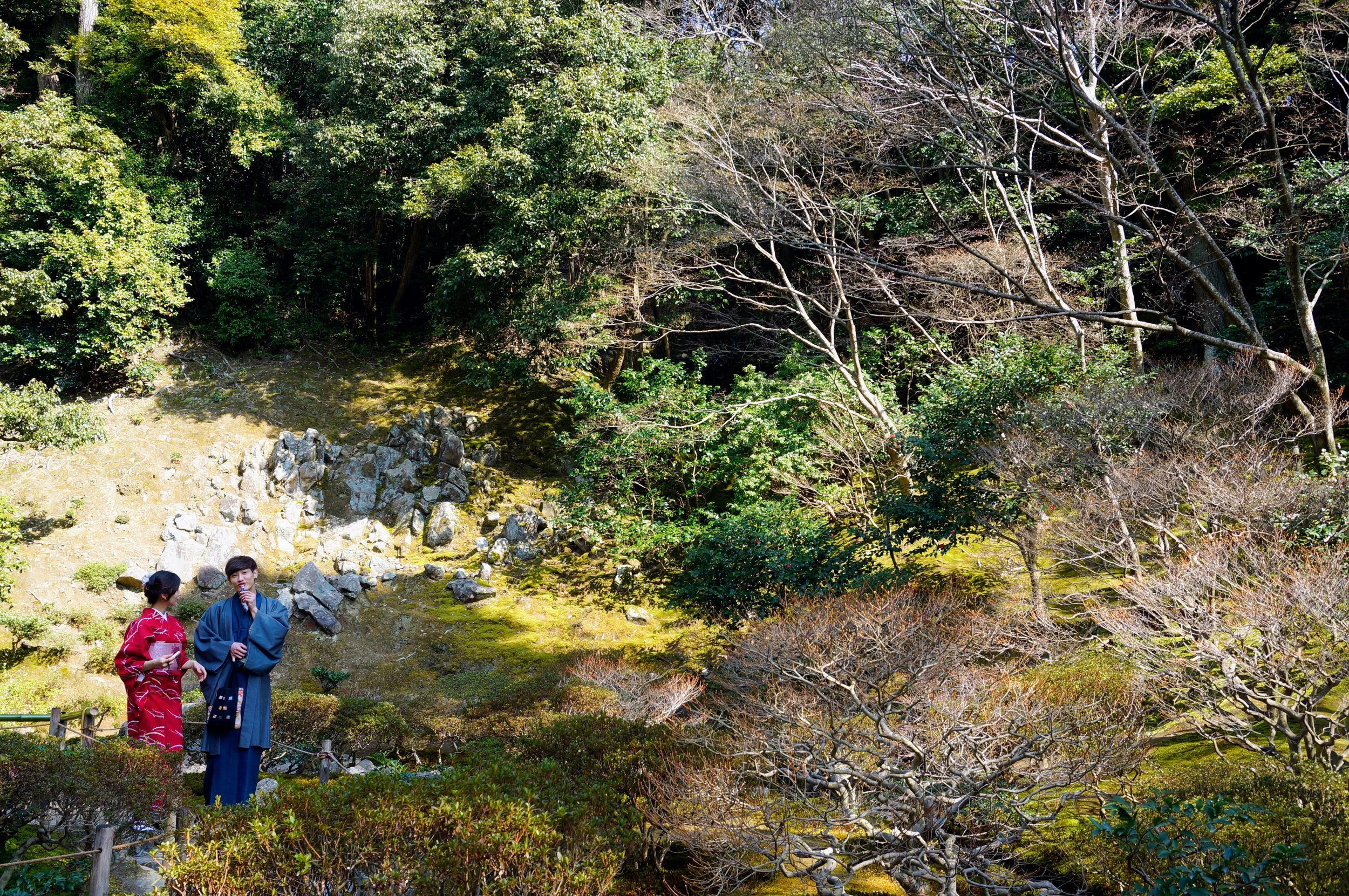 Two people in traditional Japanese clothing stand in a scenic garden with green foliage and rock formations.