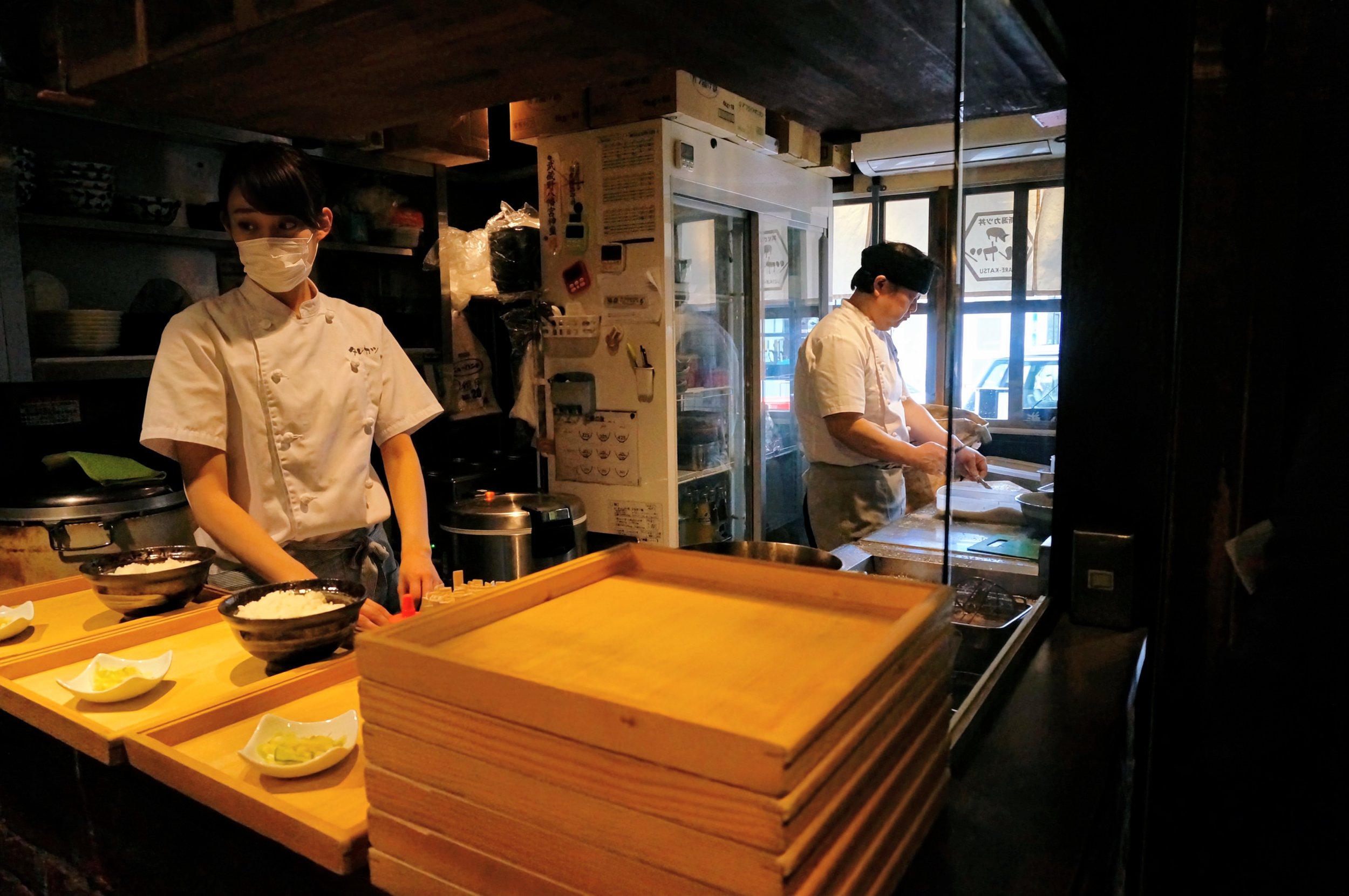 Two chefs in a Japanese restaurant kitchen, one preparing food, the other standing near trays and bowls.