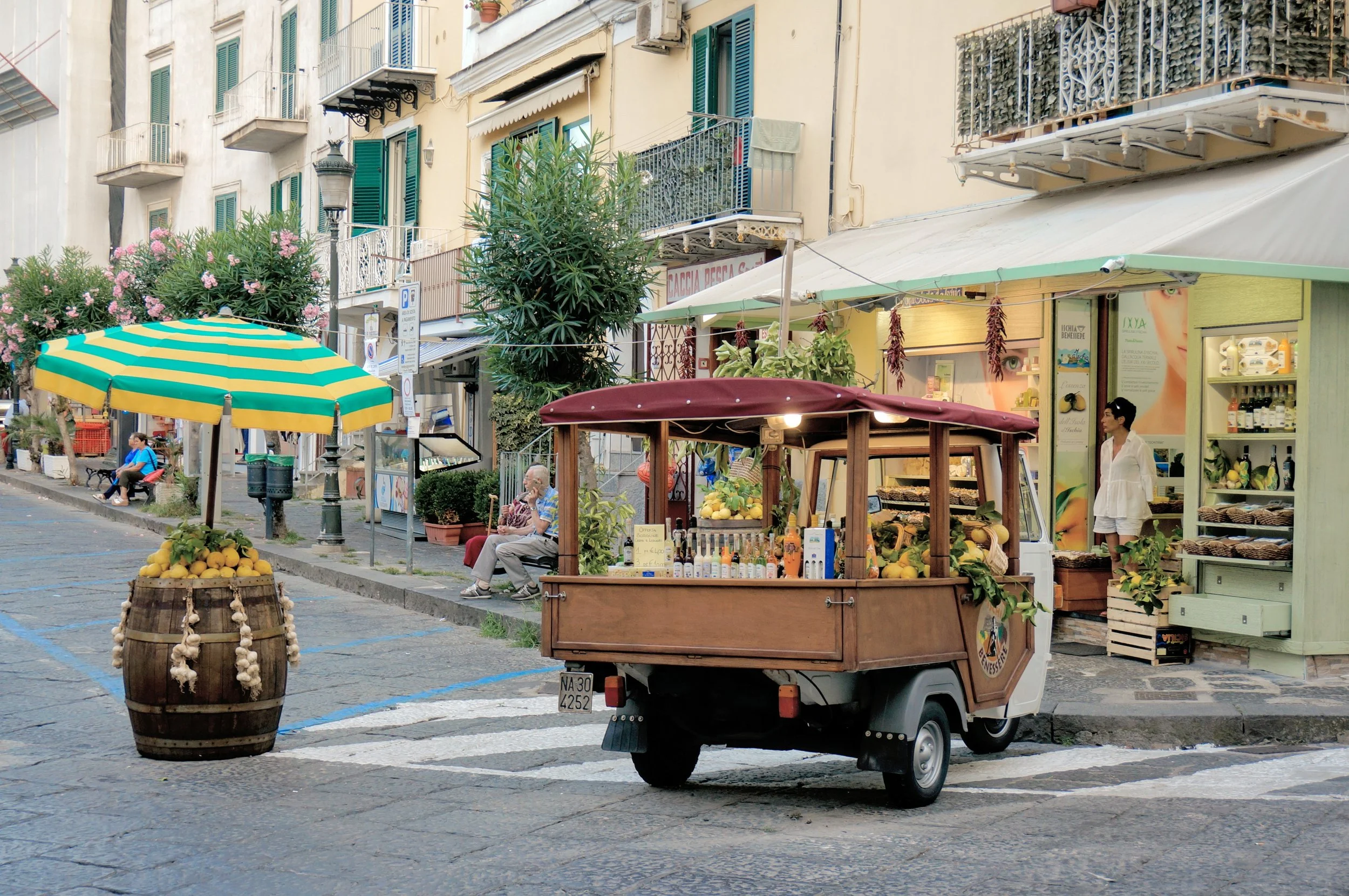 Street scene in Italy with a small fruit and beverage cart, colorful umbrella, and storefronts. People are sitting and standing nearby.