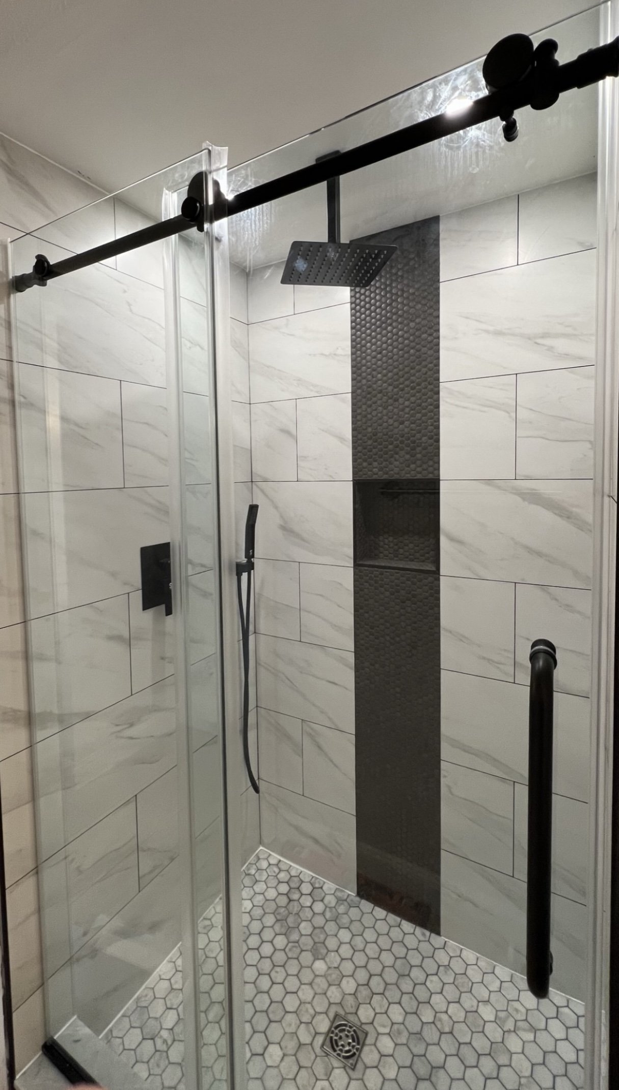 Modern shower with sliding glass doors, white marble tiles, hexagonal floor tiles, and black shower fixtures.