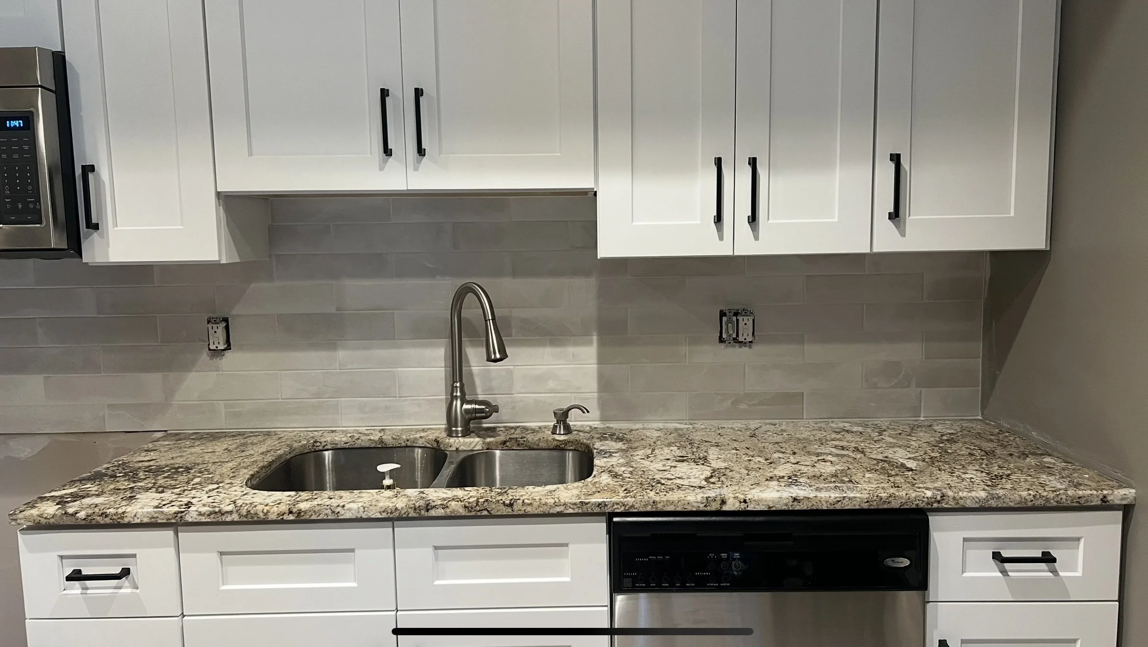 Modern kitchen with white cabinets, stainless steel sink, black hardware, granite countertop, beige tile backsplash, and built-in microwave and dishwasher.