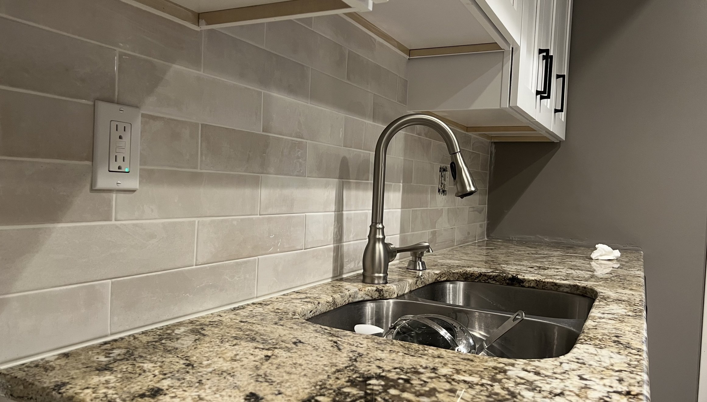 Modern kitchen counter with granite countertop, stainless steel faucet, sink, gray tile backsplash, GFCI outlet, and white cabinetry.