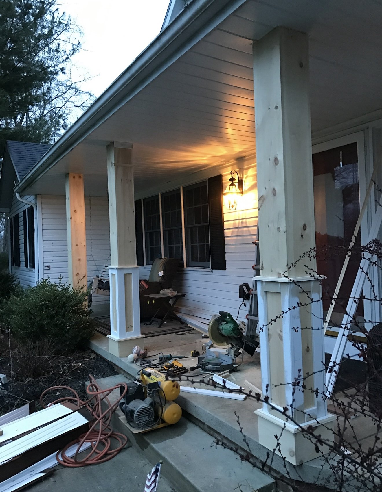 Porch with construction materials and tools, wooden columns, and a lit outdoor lamp.