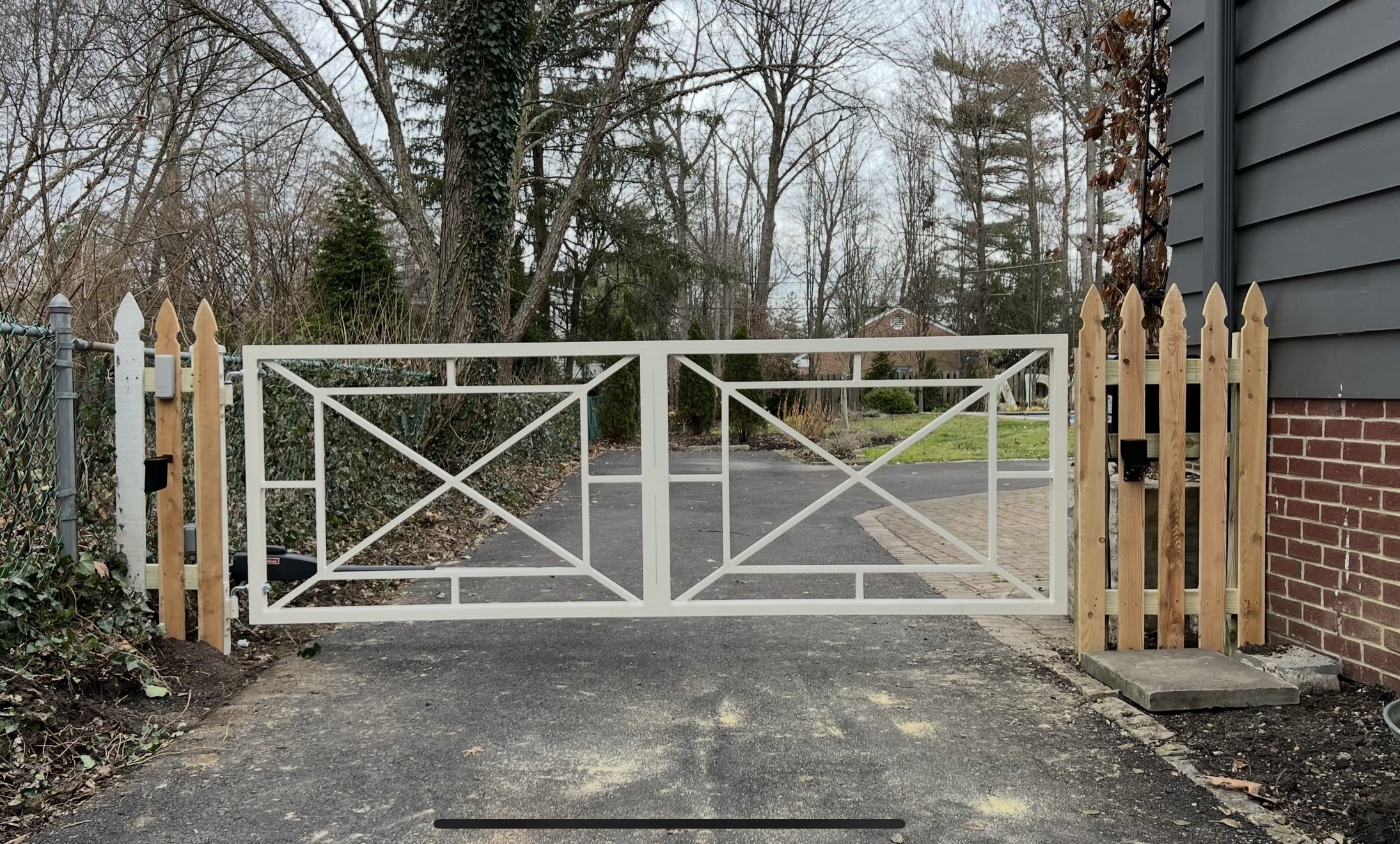 White metal gate in a driveway with geometric design and surrounding wooden and chain-link fences.