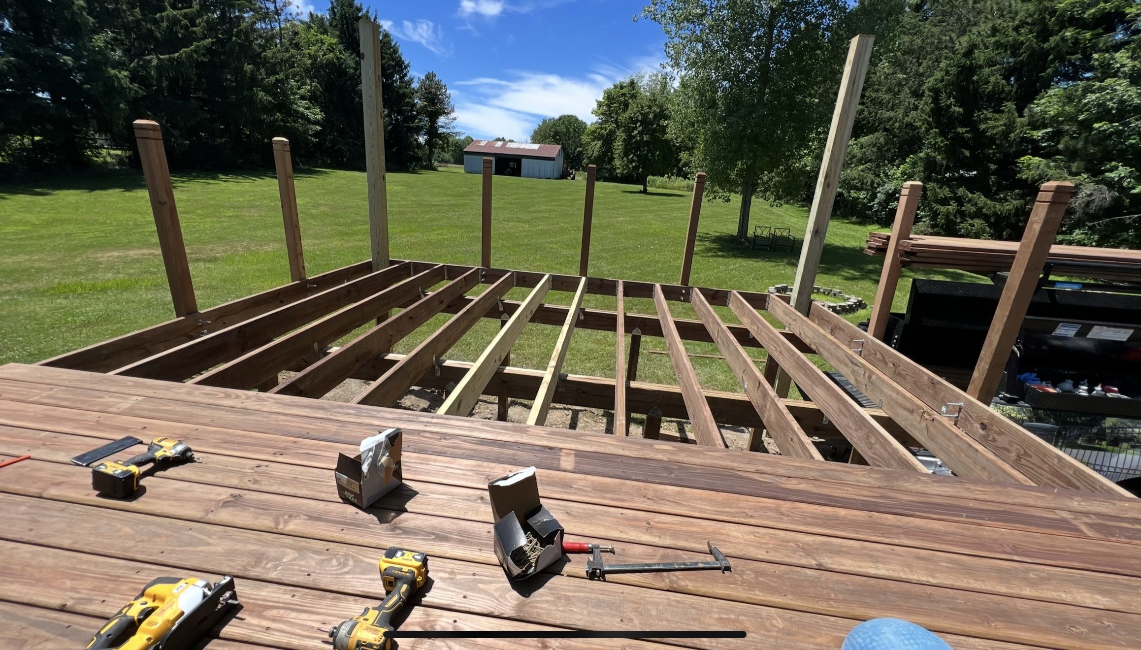 Partially constructed wooden deck with power tools and building materials on top, set in a grassy backyard with trees and a small structure in the distance.