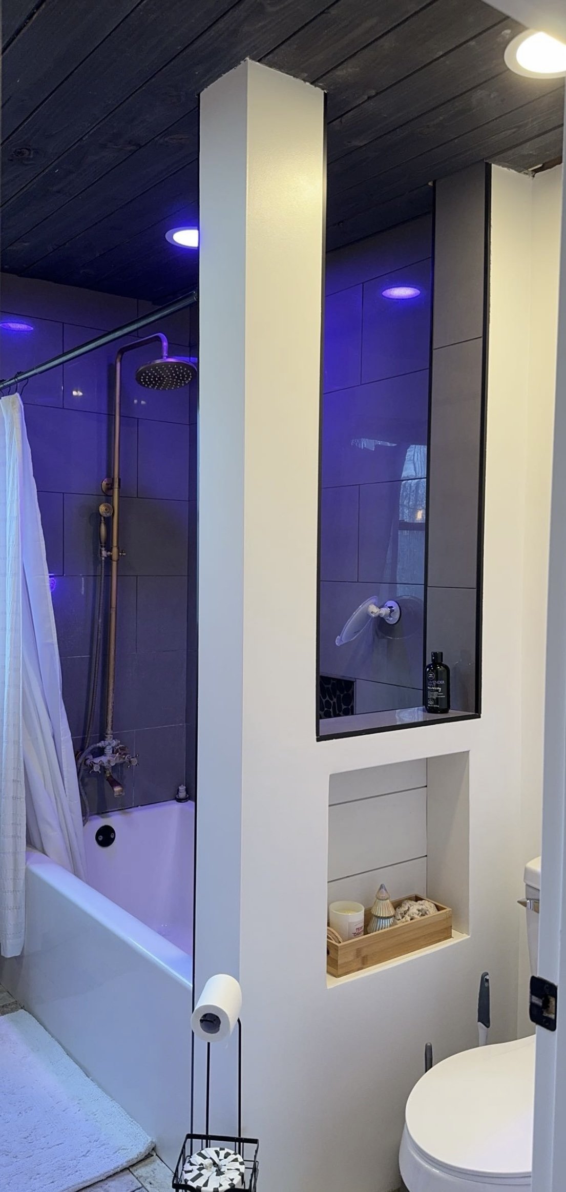 Modern bathroom with black ceiling, LED lighting, large showerhead, and white curtain in a tiled shower area. Features a recessed shelf with decor items, toilet below with toilet paper holder, and ambient lighting.