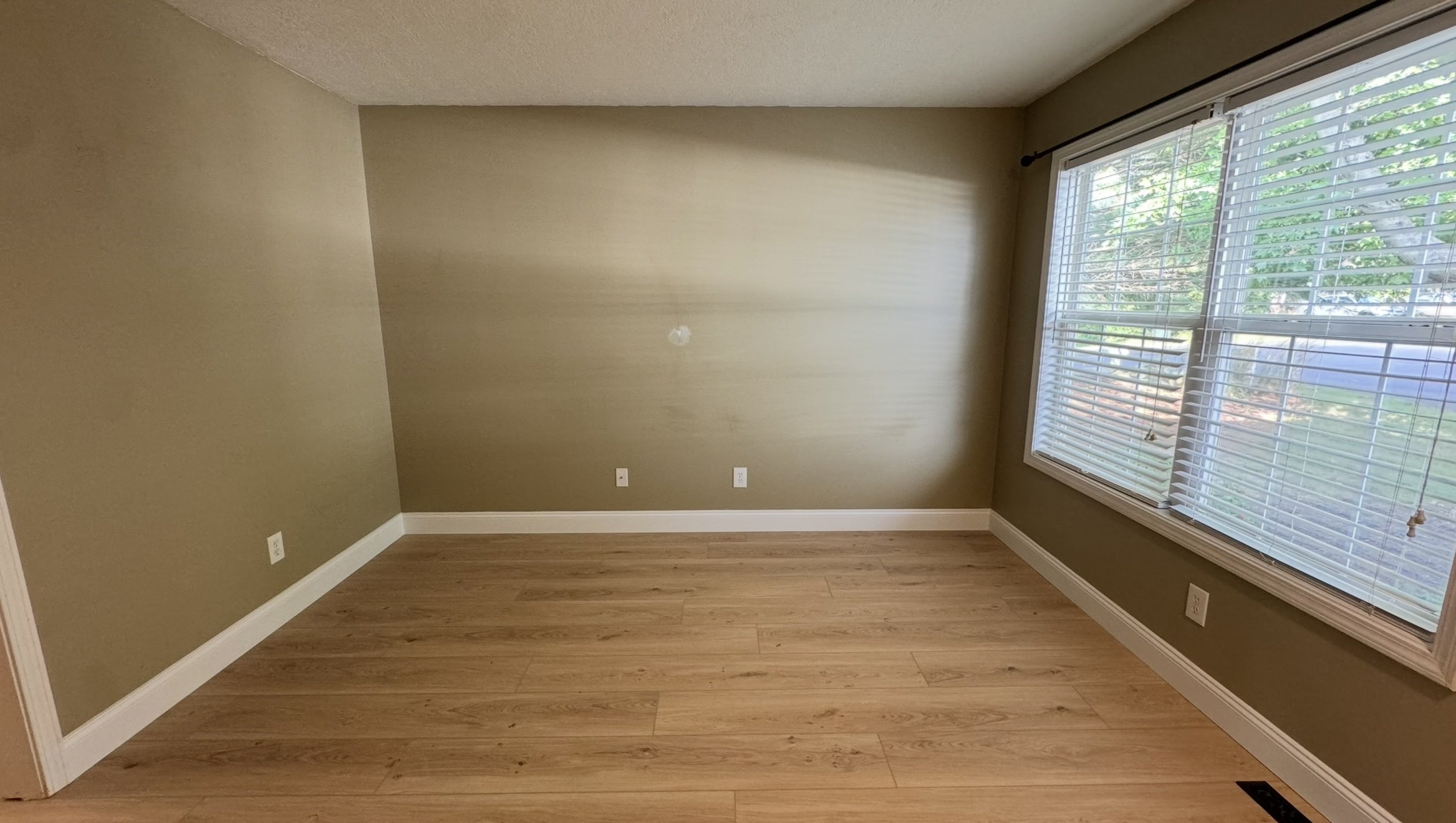 Empty room with beige walls, hardwood floor, large window with blinds, and electrical outlets on the wall.