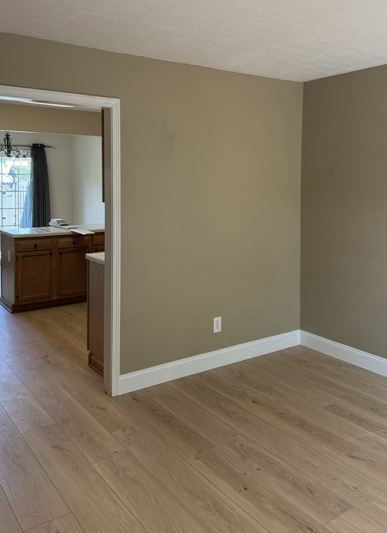 Empty room with beige walls and wood flooring, doorway leading to kitchen area with wooden cabinets and natural light.