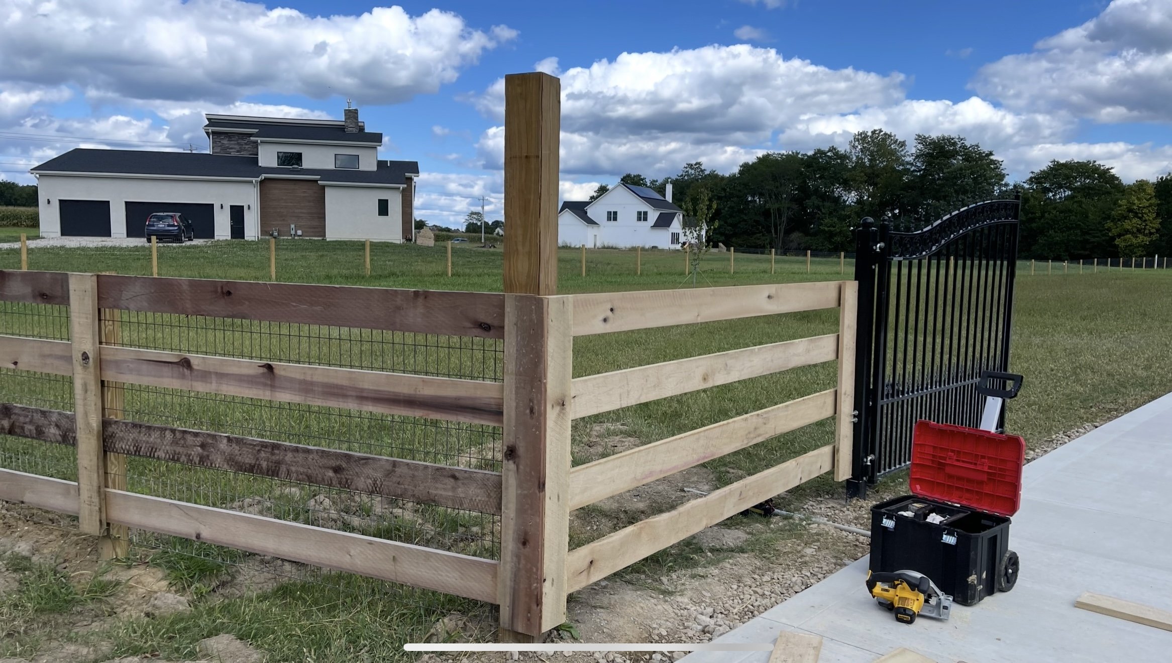 Modern house with wooden and metal fence, toolbox, and circular saw on paved driveway next to grassy area under blue sky with clouds.
