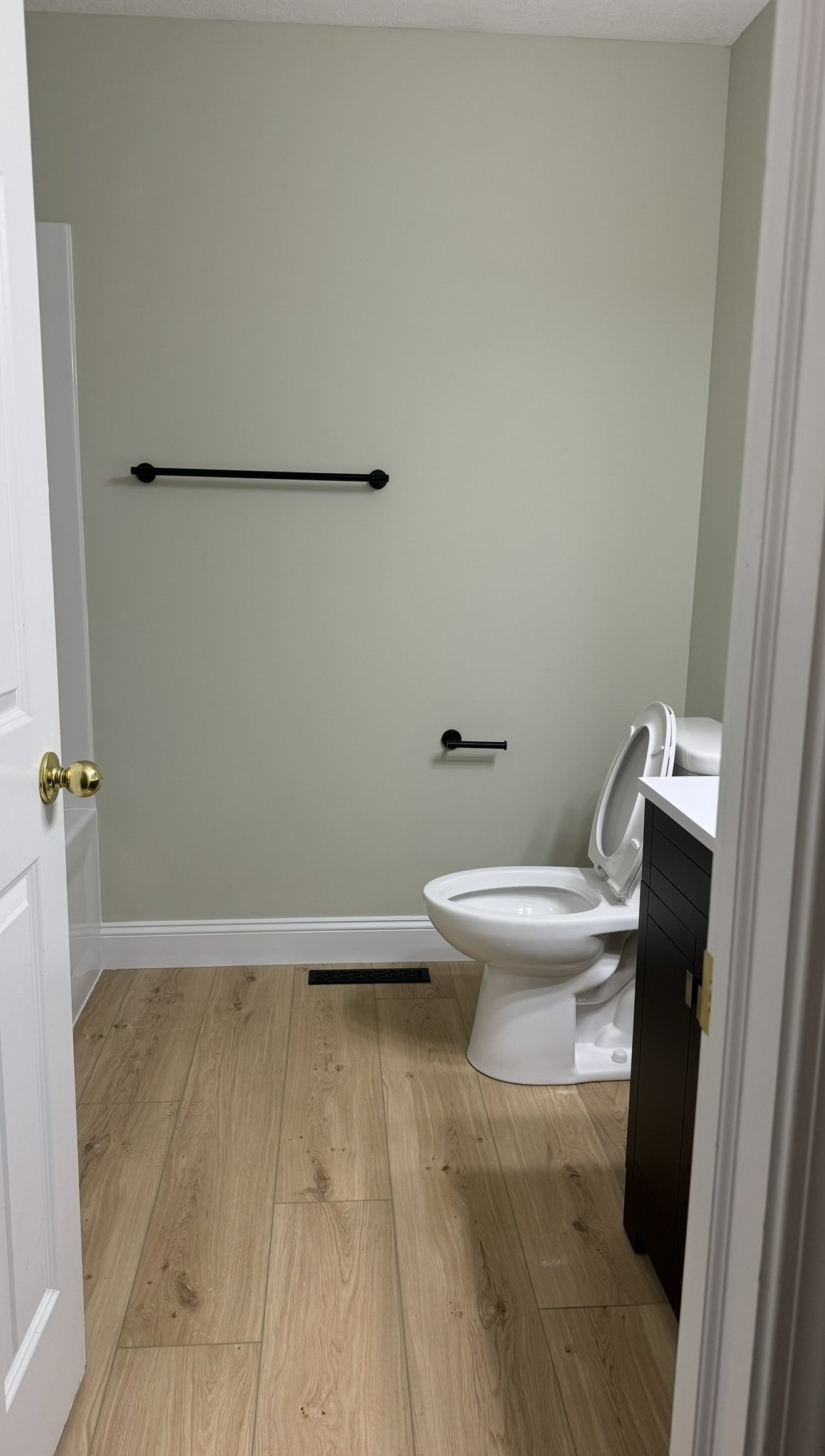 Minimalist bathroom with light wood flooring, a white toilet, a dark-colored vanity, a towel bar, and a toilet paper holder on a neutral wall.