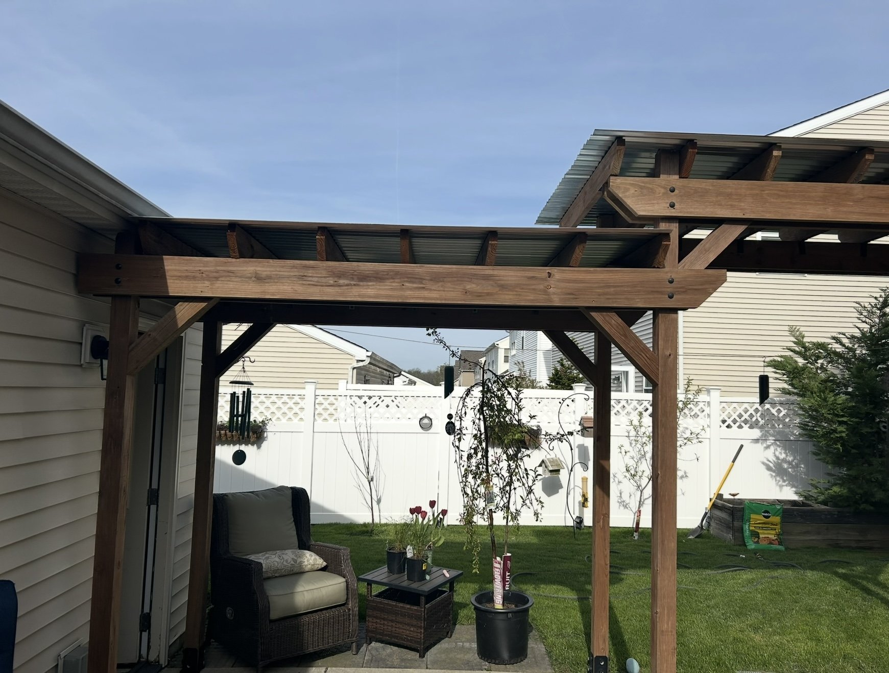 Wooden pergola over patio with wicker chair and small table in a backyard.
