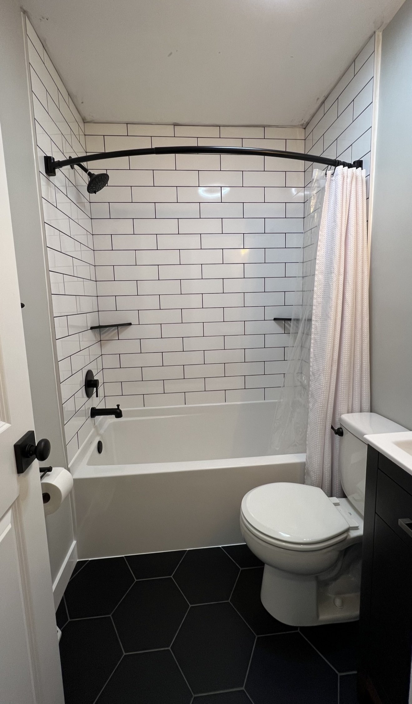 Bathroom with white subway tile, white bathtub, black shower fixtures, curved shower rod, white curtain, dark hexagonal floor tiles, toilet, white walls, and a vanity.