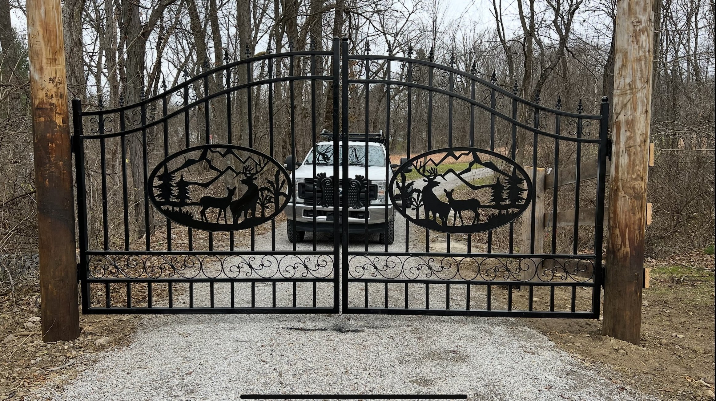 Black wrought iron gate with deer and mountain cutouts, closed across a gravel driveway, with a vehicle visible in the background, surrounded by leafless trees.