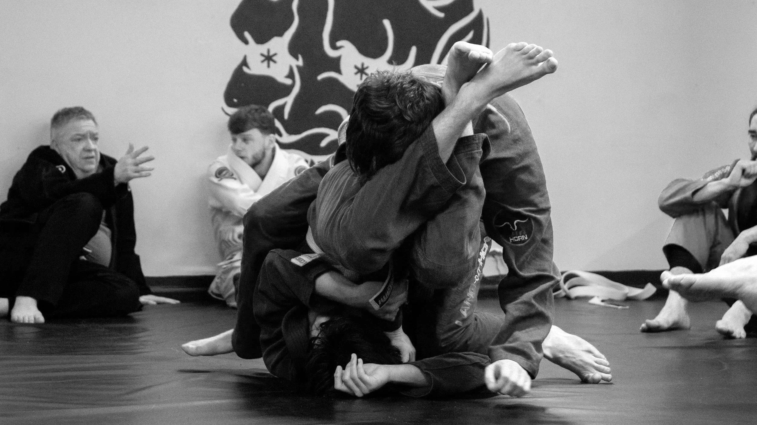 A group of people practicing Brazilian Jiu-Jitsu on a mat, with one person attempting a grappling hold on another, while others observe and discuss nearby.