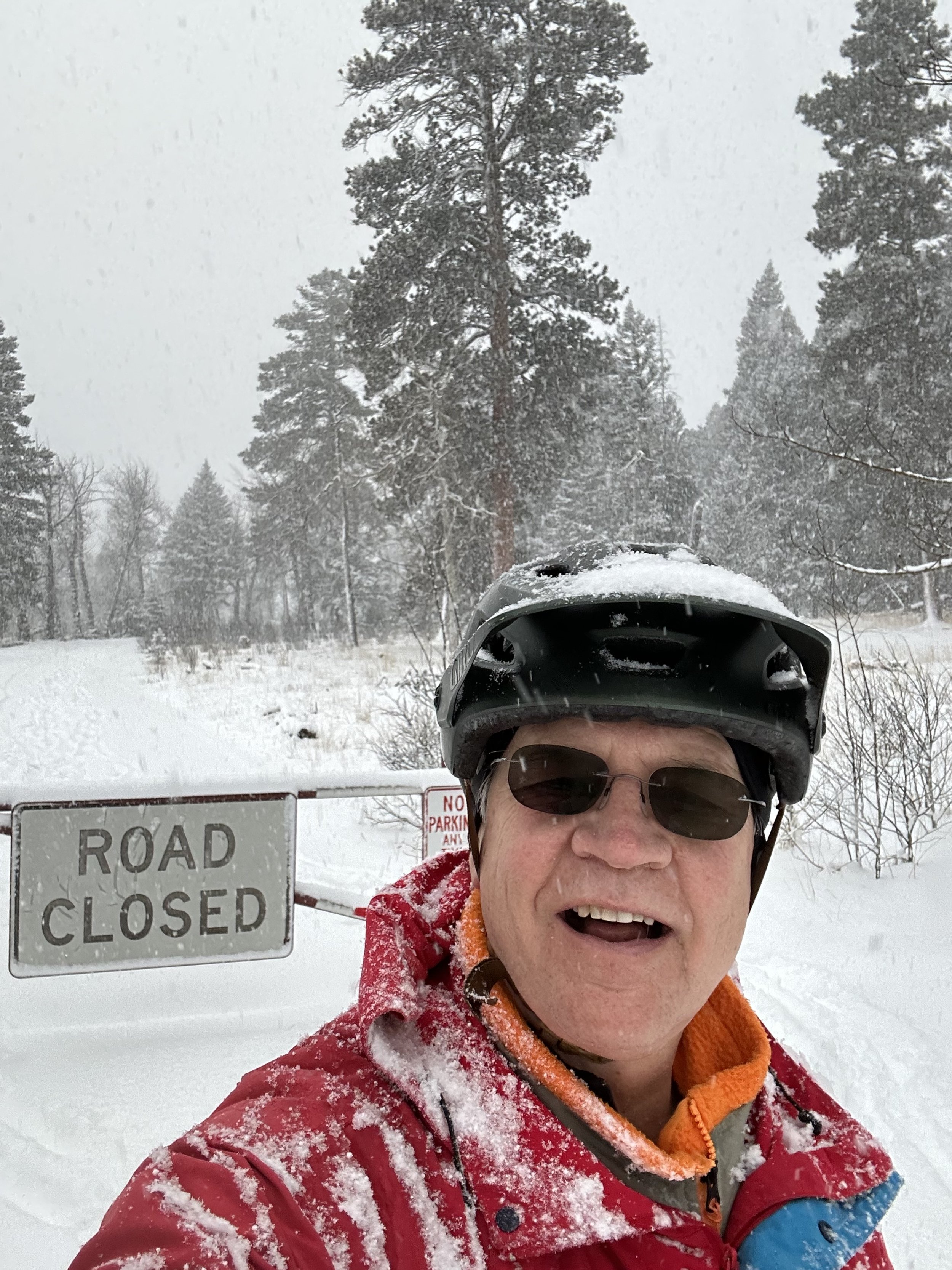 Dr. Leigh wearing a helmet and sunglasses is taking a selfie in a snowy landscape. They are dressed in a red jacket with snow on it. In the background, there is a "Road Closed" sign and snow-covered trees.