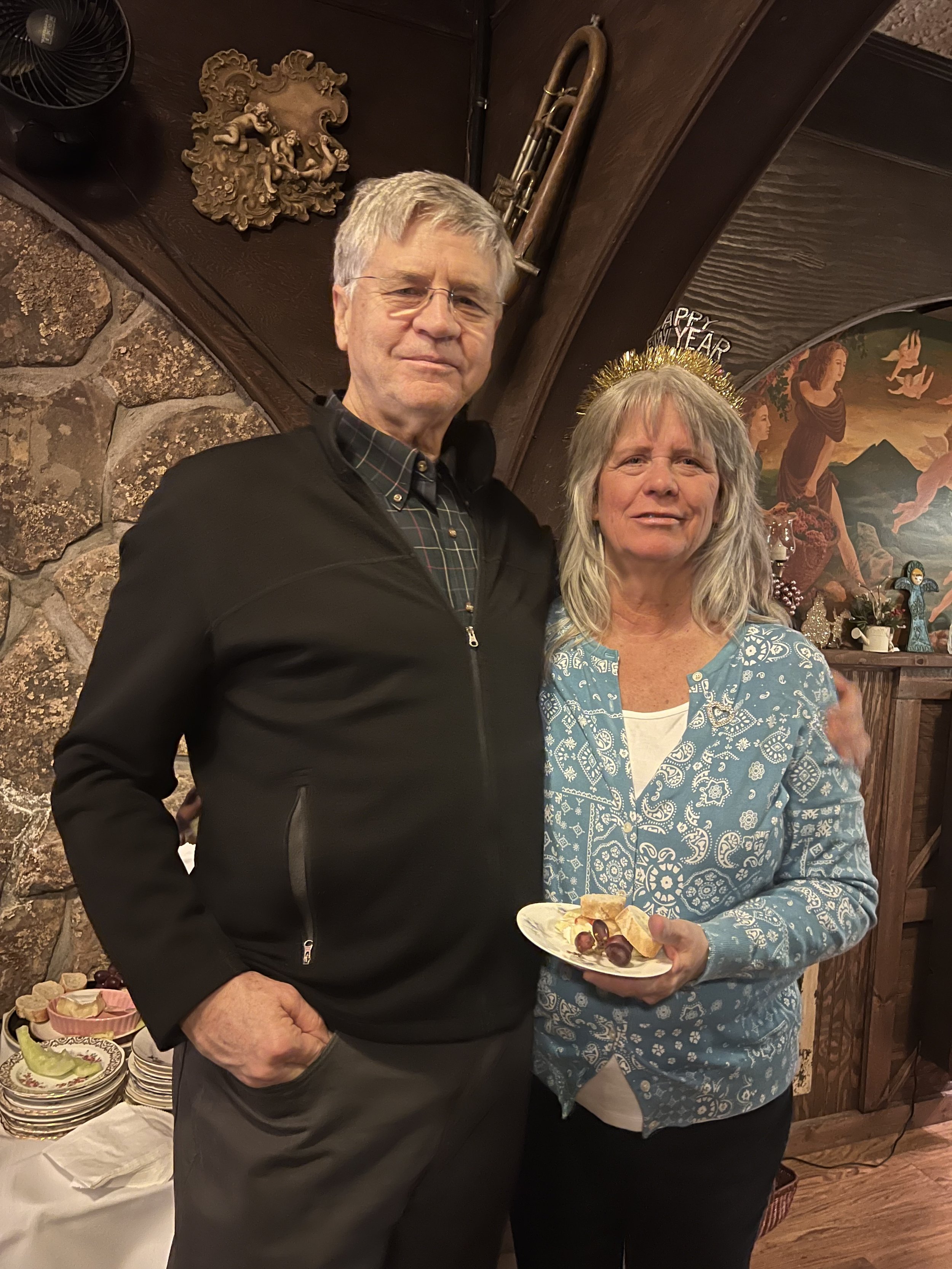Dr. Tom Leigh and his wife posing indoors, the woman holding a plate with snacks. She wears a "Happy New Year" headband. The background features decorative elements like a mural and a sculpture on a stone wall.