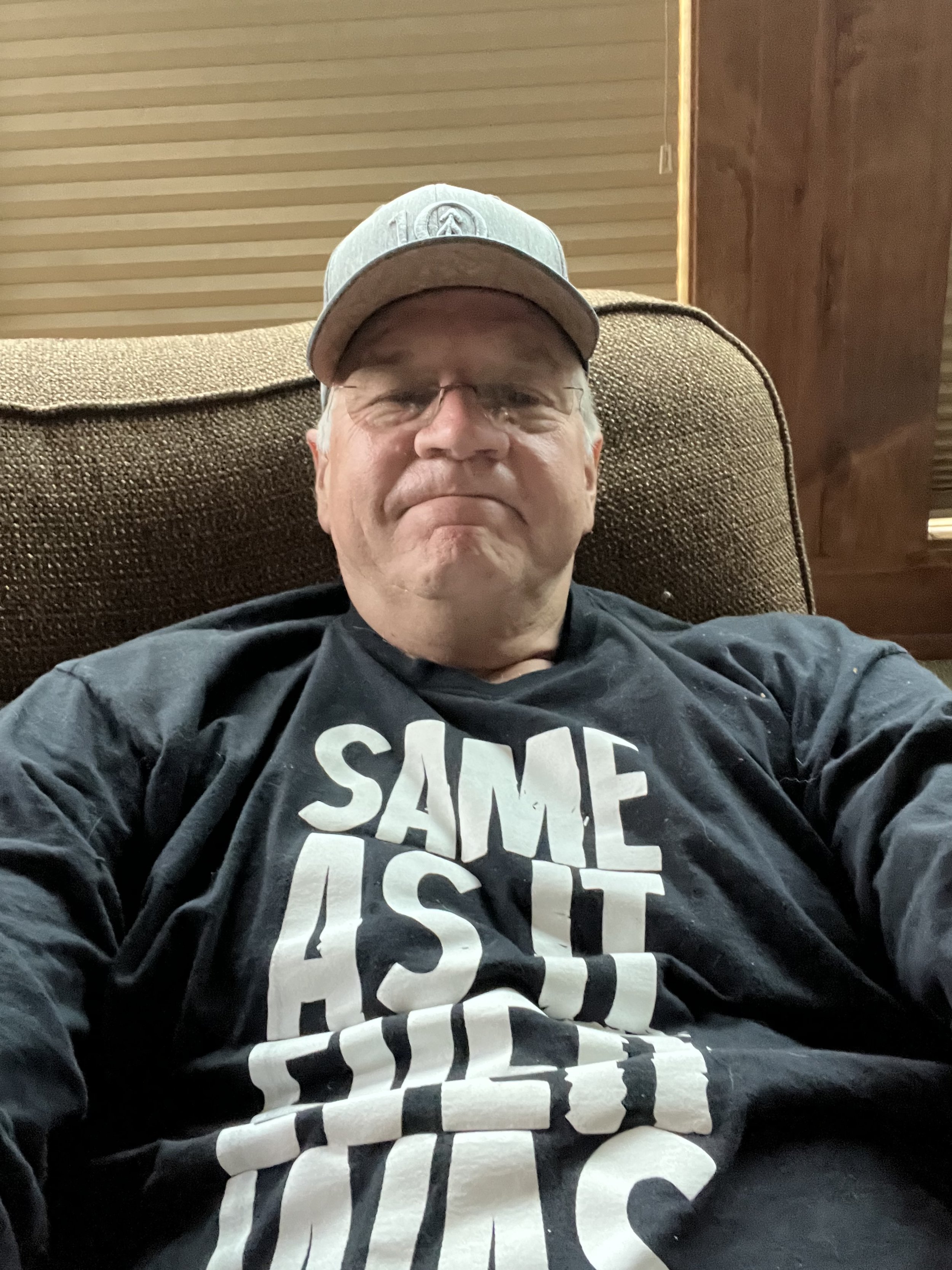 Dr. Leigh wearing a gray cap and black shirt with white text that says 'SAME AS IT EVER WAS' sitting on a brown couch.