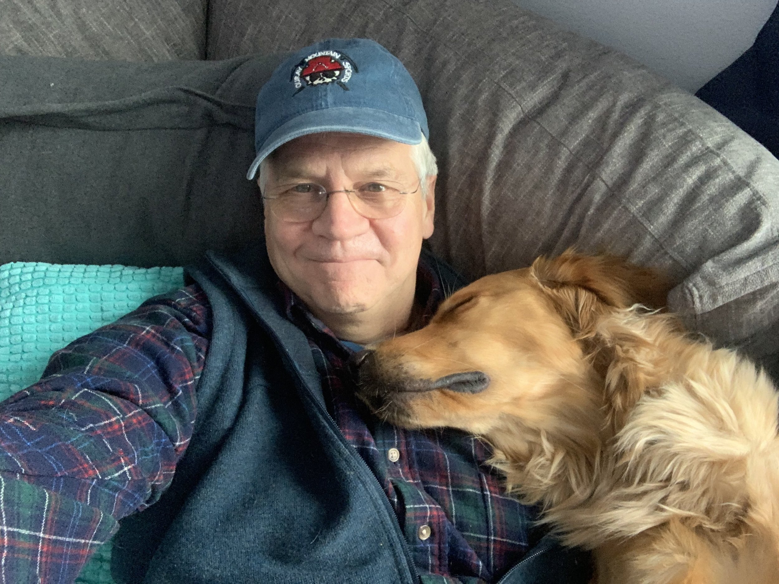 Dr. Tom Leigh wearing glasses and a baseball cap smiles while a golden retriever sleeps on his chest on a couch.