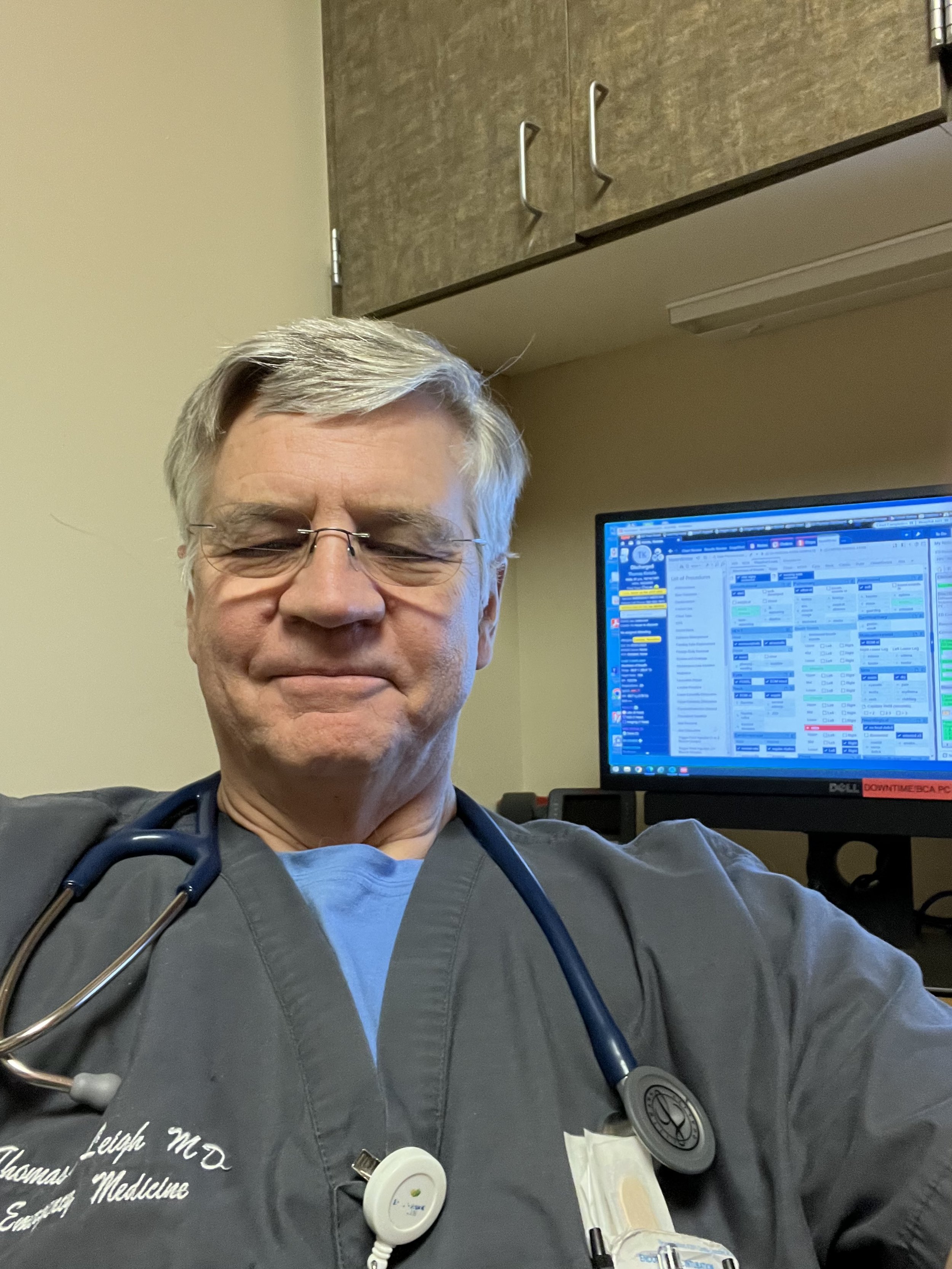 Dr. Tom Leigh wearing a stethoscope around their neck, sitting in front of a computer screen displaying medical data. The person is wearing glasses and a dark medical scrub top.