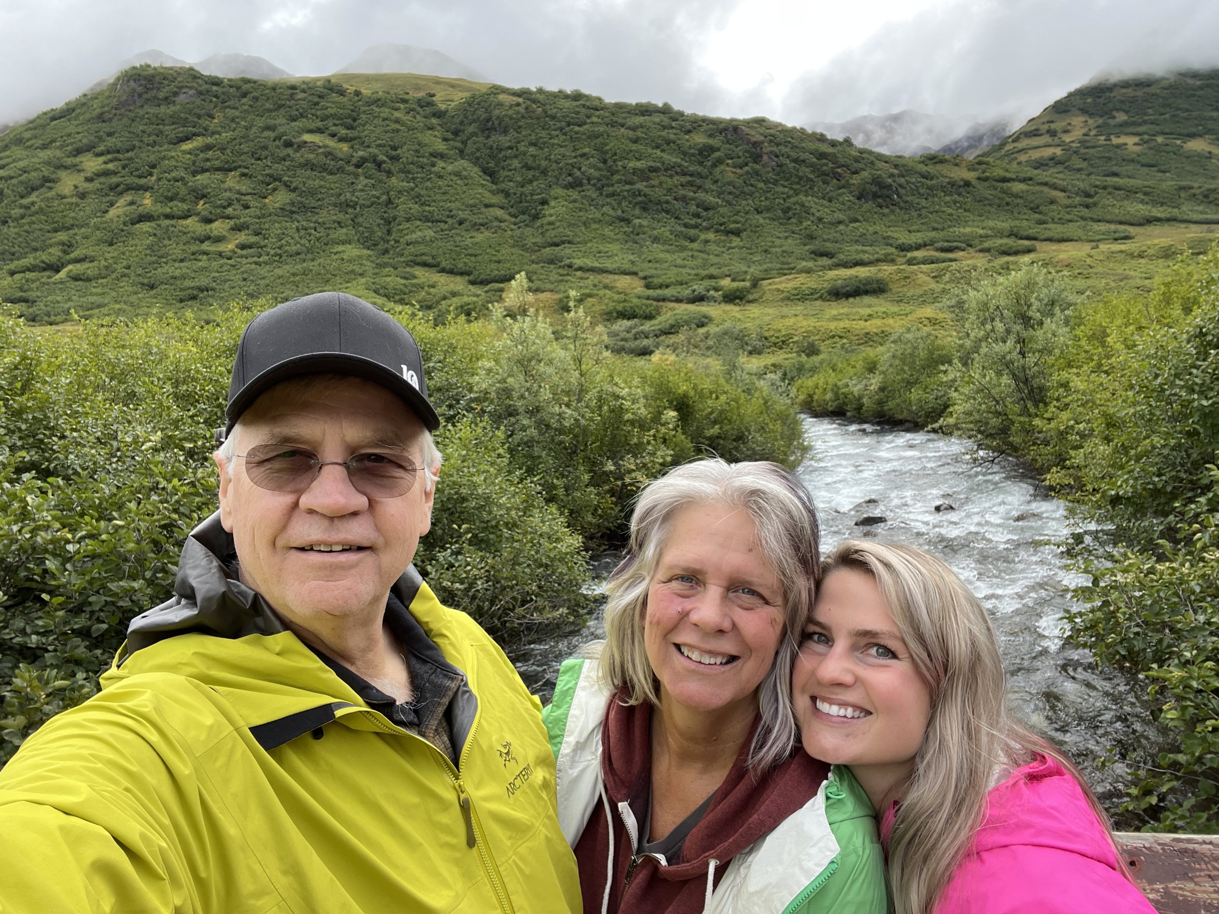 Dr. Leigh on a trip people smiling in front of a scenic landscape with a river, lush green hills, and a cloudy sky.