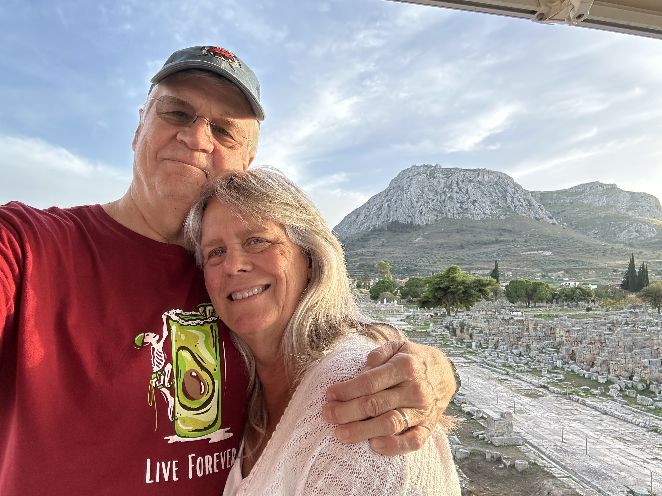 Dr. Thomas Leigh and his wife standing close together and smiling, with an ancient ruins site and a mountainous landscape in the background, under a partly cloudy sky.