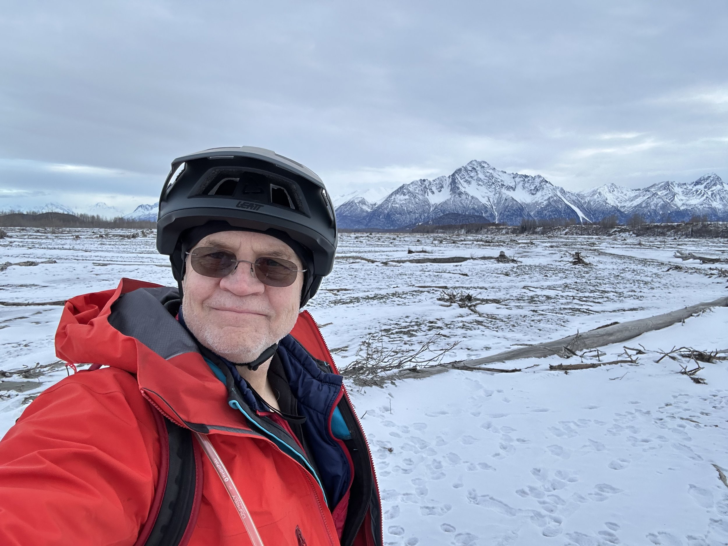 Dr. Leigh Man in a red jacket with a helmet standing in a snowy landscape with mountains in the background.