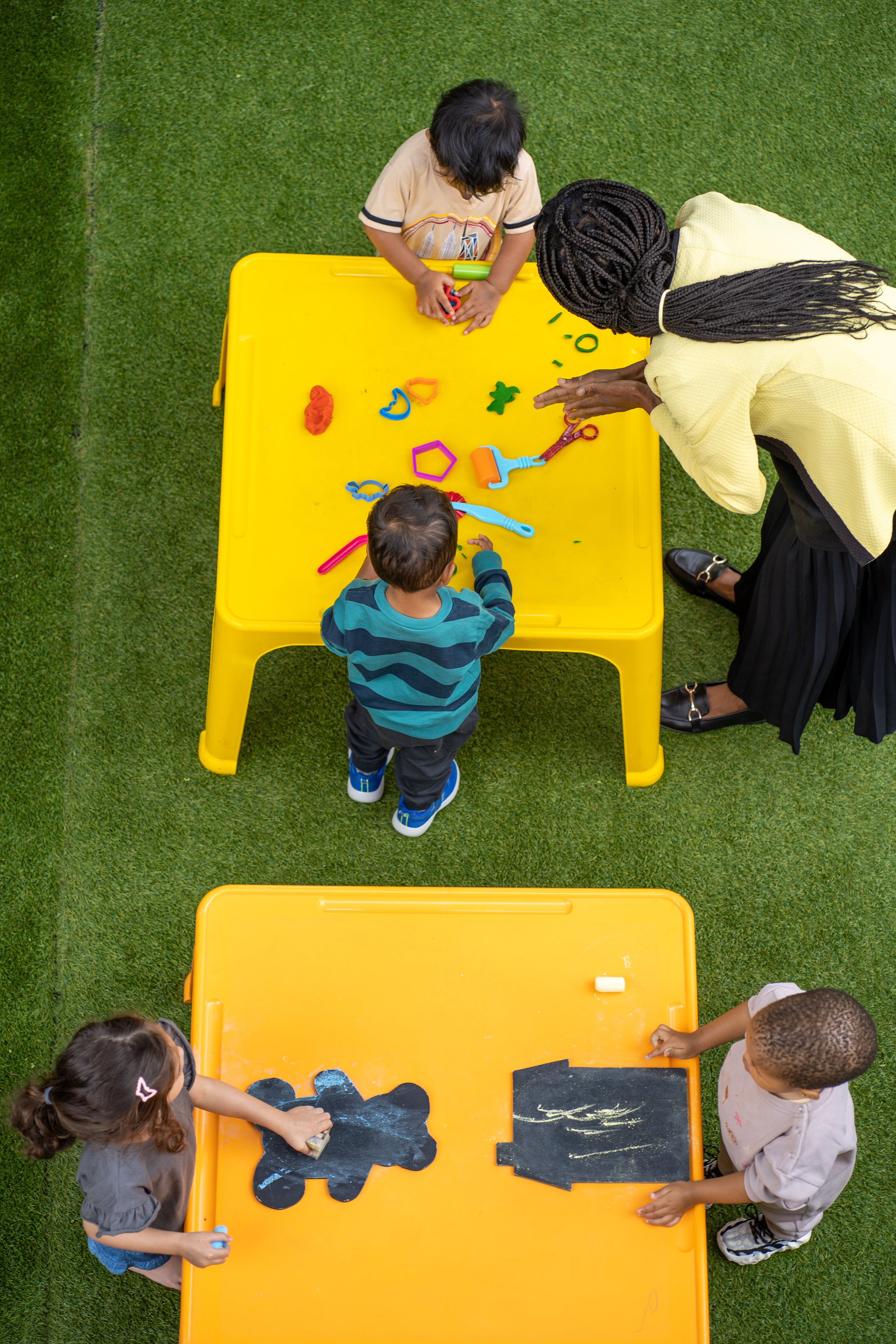 Two children and an adult playing with toys at yellow tables outdoors on grass.