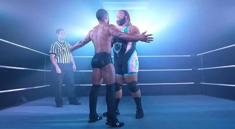Two male wrestlers face off in a wrestling ring, with a referee observing in the background.