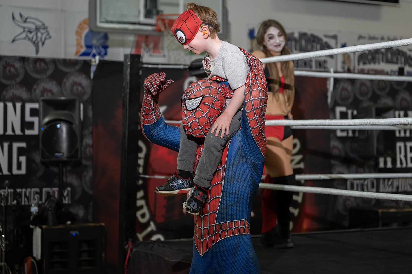 A person dressed as Spider-Man holding a child on their shoulders during a Halloween event at a wrestling ring, with a person in a Halloween costume in the background.