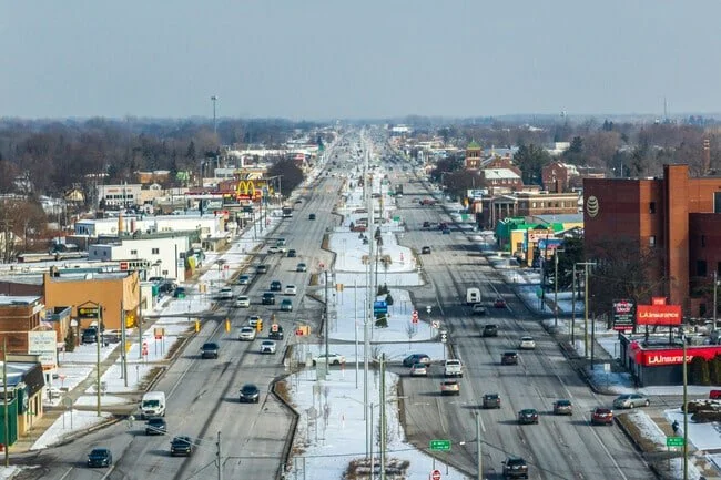 A busy four-lane road in a commercial area with cars and trucks, surrounded by shops and restaurants, with snow on the ground and a cloudy sky.
